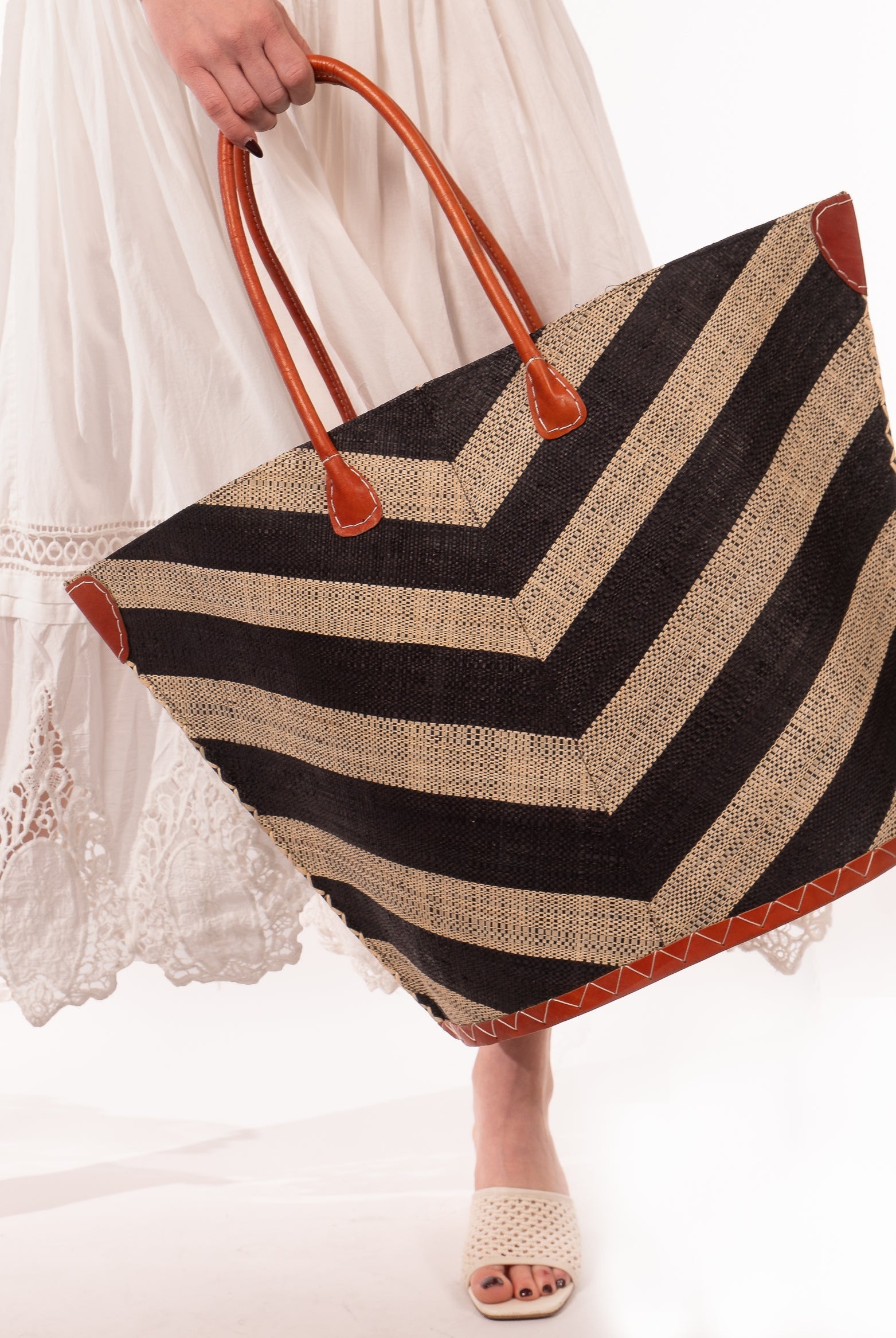 Model wearing Santa Cruz Wide Stripe Chevron Large Straw Tote Bag handmade loomed raffia palm fiber in a two tone diagonal even width bands of color in a wide stripe chevron pattern of black and natural straw color on the front and a matching solid hue on the back with leather accents and handles large shoulder bag - Shebobo