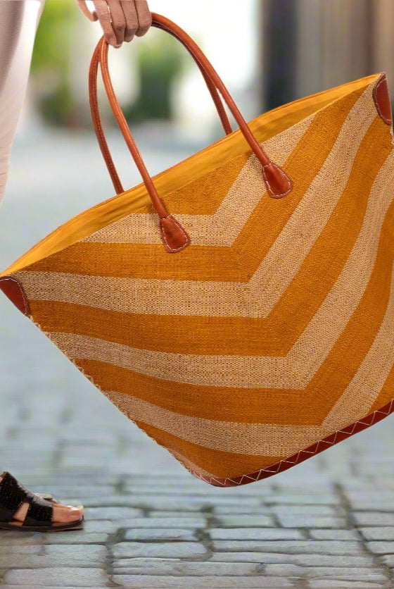 Model wearing Santa Cruz Wide Stripe Chevron Large Straw Tote Bag handmade loomed raffia palm fiber in a two tone diagonal even width bands of color in a wide stripe chevron pattern of saffron orange/yellow and natural straw color on the front and a matching solid hue on the back with leather accents and handles large shoulder bag - Shebobo
