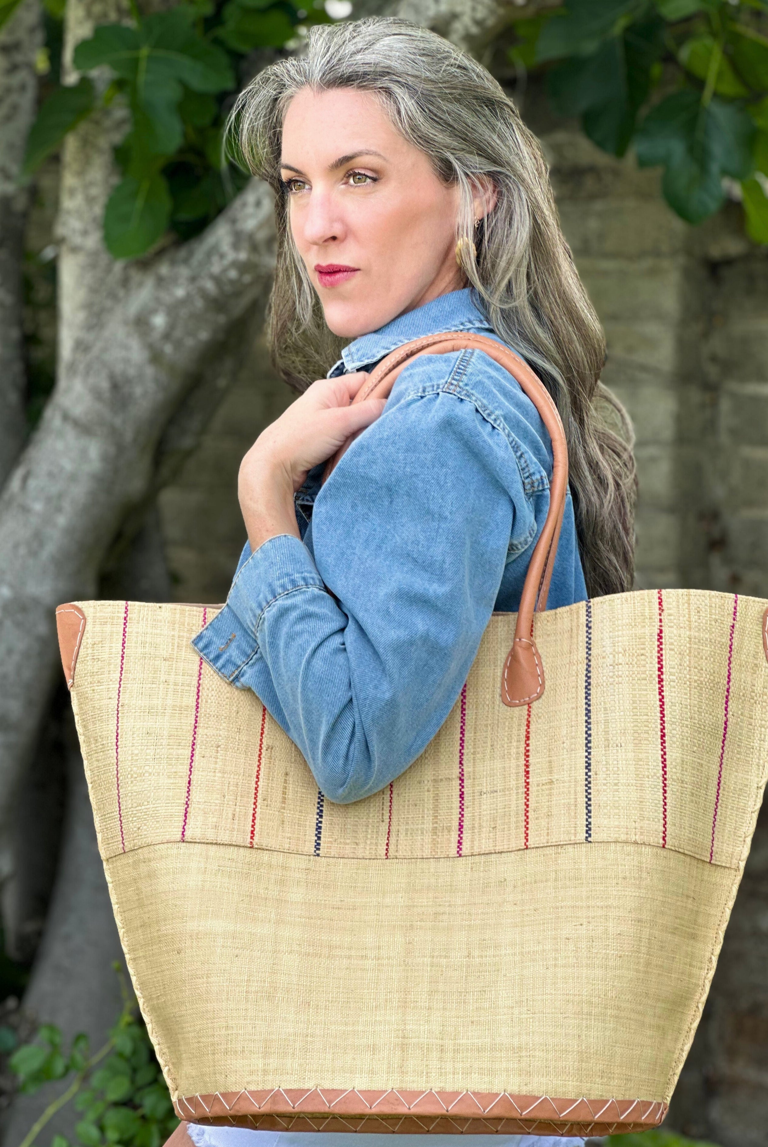 Model wearing Santa Cruz Two Tone Pinstripes Multi Large Straw Tote Bag handmade loomed raffia in a wide stripe of natural straw color with multicolor thin stripes of fuchsia pink, coral red/orange, and navy blue in a vertical pinstripe pattern on the top half and solid natural on the bottom half handbag with leather handles and details beach bag - Shebobo