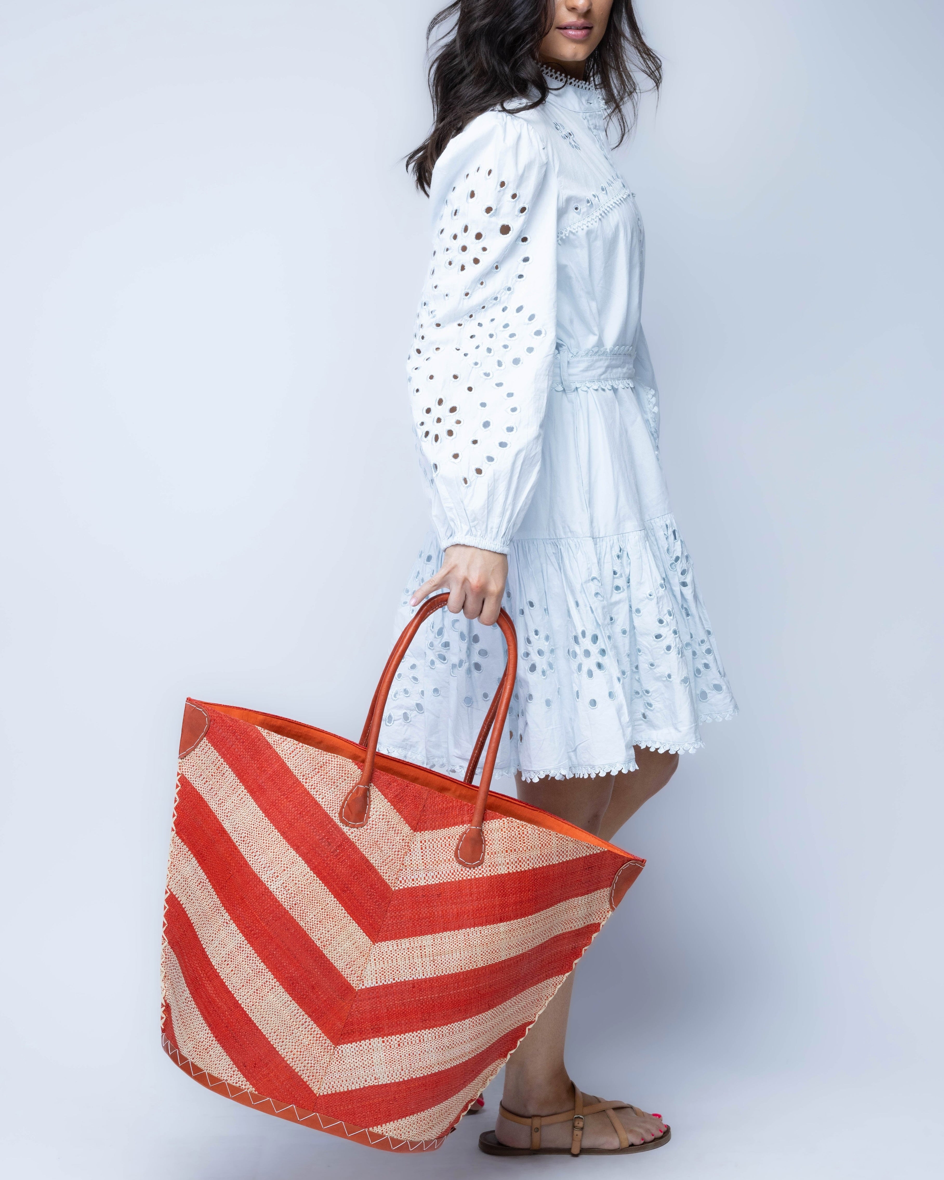 Model wearing Santa Cruz Wide Stripe Chevron Large Straw Tote Bag handmade loomed raffia palm fiber in a two tone diagonal even width bands of color in a wide stripe chevron pattern of coral orange/red and natural straw color on the front and a matching solid hue on the back with leather accents and handles large shoulder bag - Shebobo