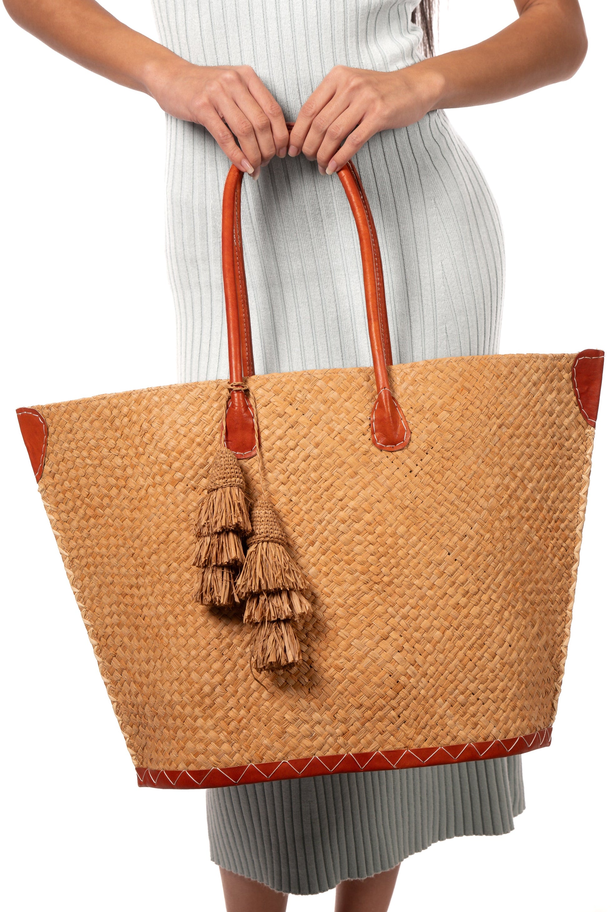 Model wearing Santa Ynez Cappuccino Woven Small Straw Tote Bag with Tassel Charm Embellishment handmade woven raffia palm fiber in a solid hue of cappuccino tea/light/brown creating a subtle wide cross weave texture with matching crochet topped multi tuft tassel embellishment and leather handles shoulder bag beach bag - Shebobo