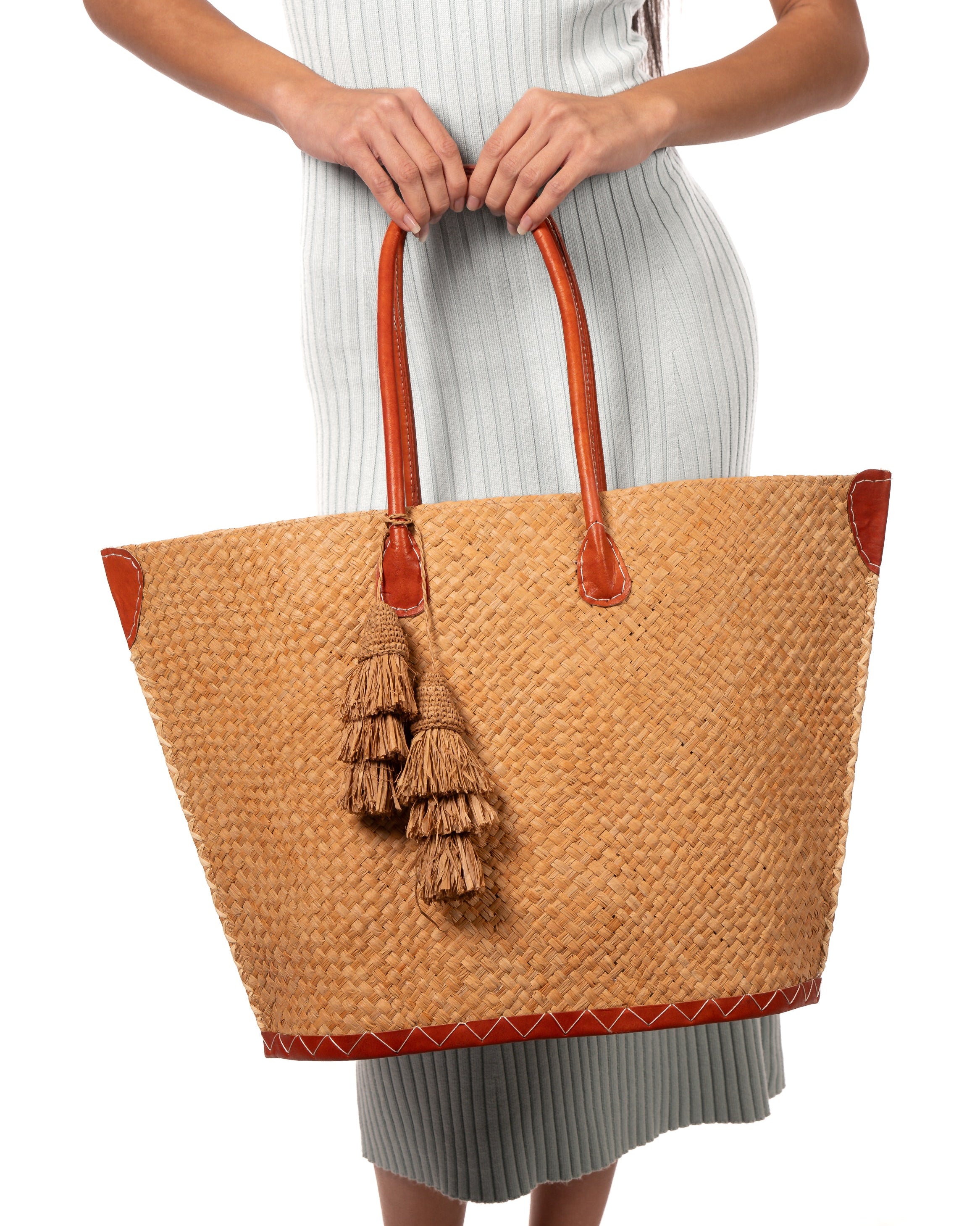 Model wearing Santa Ynez Cappuccino Woven Small Straw Tote Bag with Tassel Charm Embellishment handmade woven raffia palm fiber in a solid hue of cappuccino tea/light/brown creating a subtle wide cross weave texture with matching crochet topped multi tuft tassel embellishment and leather handles shoulder bag beach bag - Shebobo