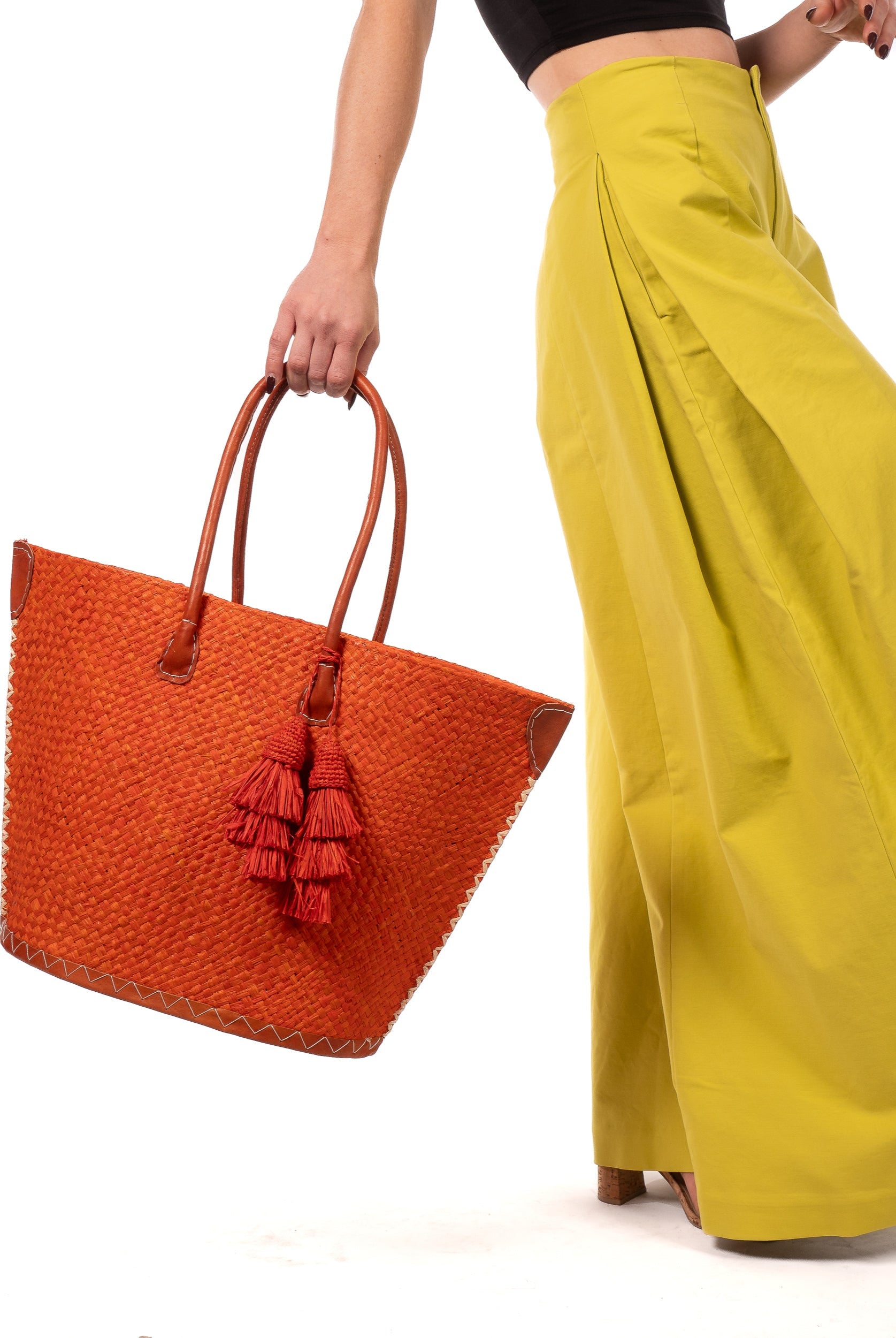 Model wearing Santa Ynez Woven Small Straw Tote Bag with Tassel Charm Embellishment handmade woven raffia palm fiber in a solid hue of coral orange/red with matching tassel tie charm embellishment and leather handles beach bag - Shebobo