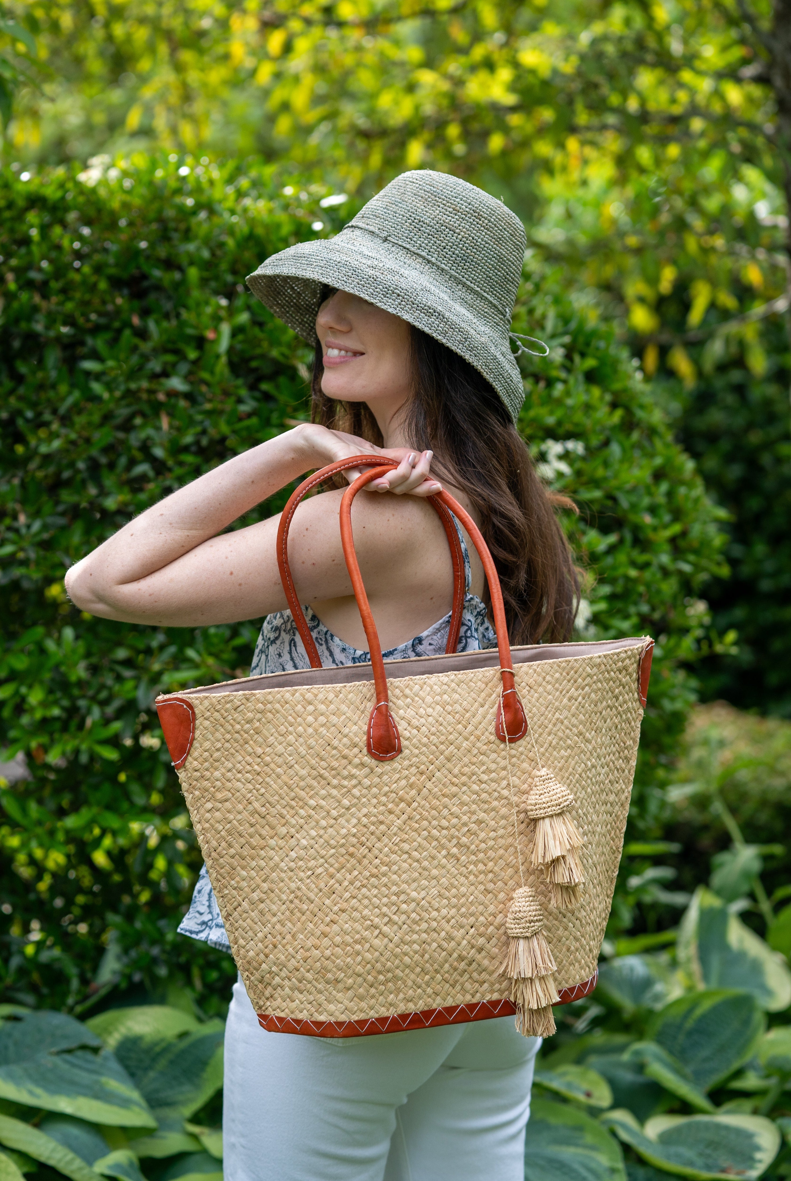 Model wearing Santa Ynez Woven Small Straw Tote Bag with Tassel Charm Embellishment handmade woven raffia palm fiber in a solid hue of natural straw color with matching tassel tie charm embellishment and leather handles beach bag - Shebobo (with 3" Brim Leor Crochet Straw Sun Hats)