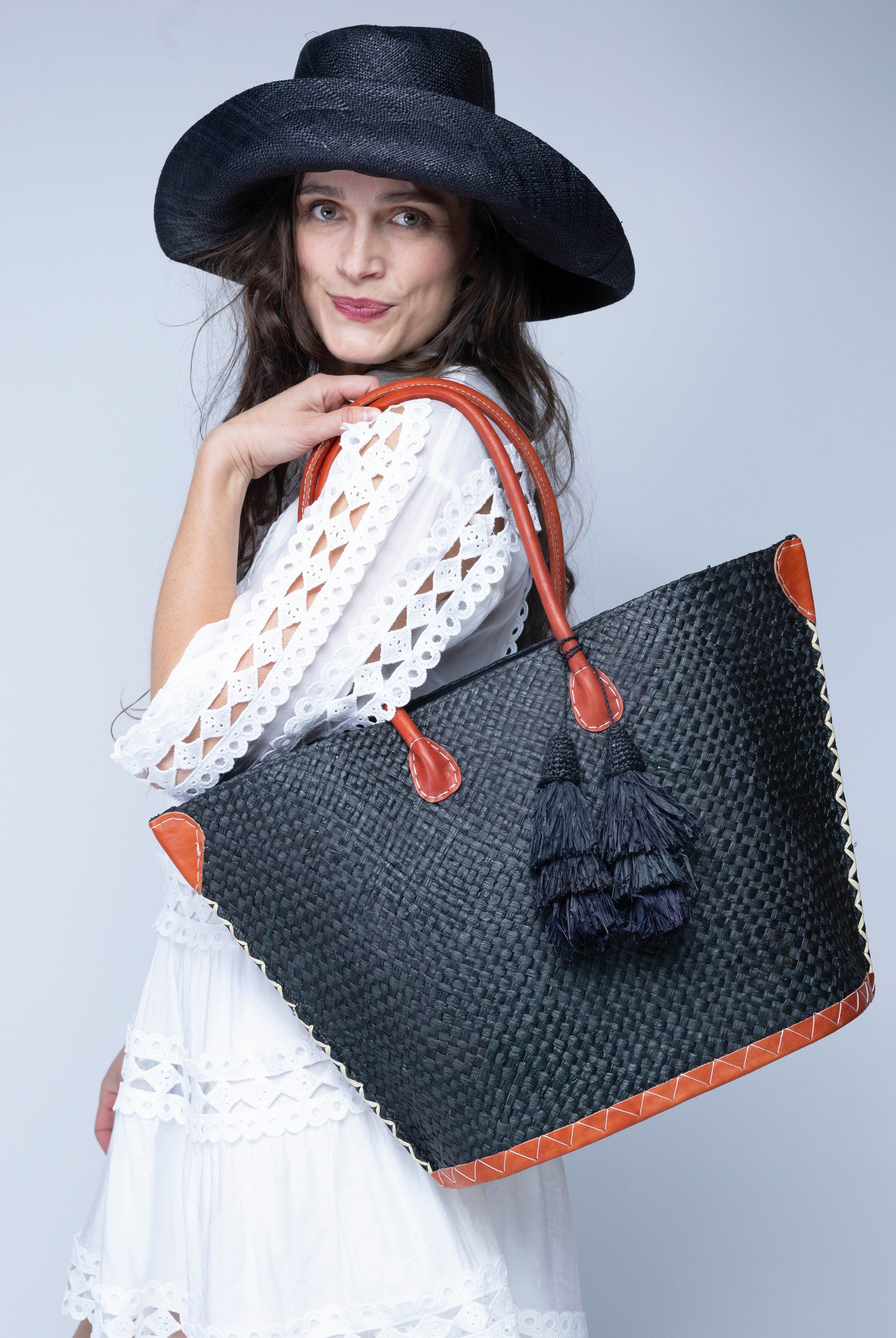 Model wearing Santa Ynez Woven Small Straw Tote Bag with Tassel Charm Embellishment handmade woven raffia palm fiber in a solid hue of black with matching tassel tie charm embellishment and leather handles beach bag - Shebobo (with Monica 5" Wide Brim Packable Straw Sun Hat)
