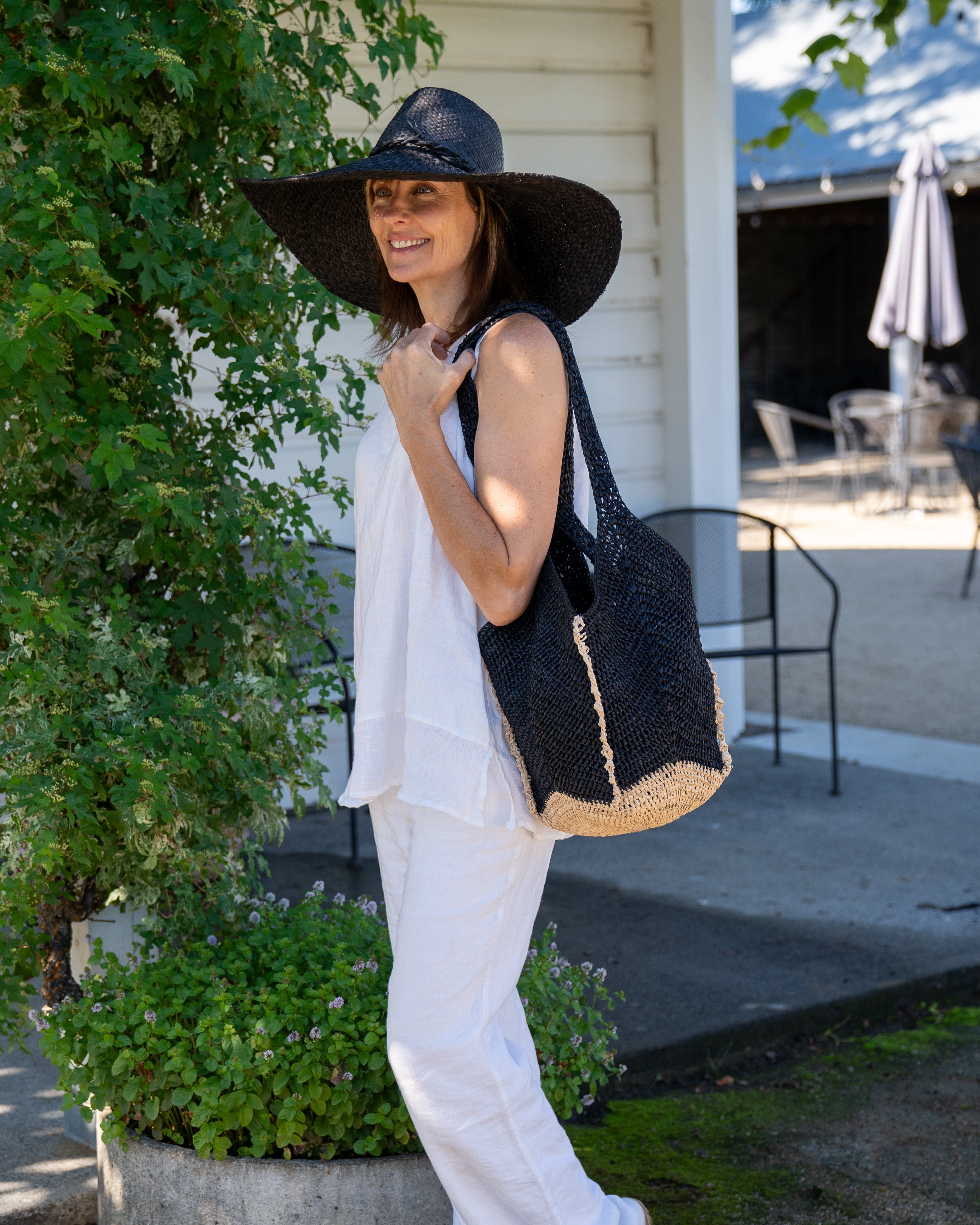 Model wearing Sea See Black Straw Shoulder Bag handmade woven raffia palm fiber in a solid hue of black making a diagonal link pattern with natural straw color crochet circular bottom and vertical edging along the side panels creating a two tone multicolor handbag/beach bag/purse - Shebobo (with Natasha 7" Wide Brim Woven XL Straw Sun Hats)