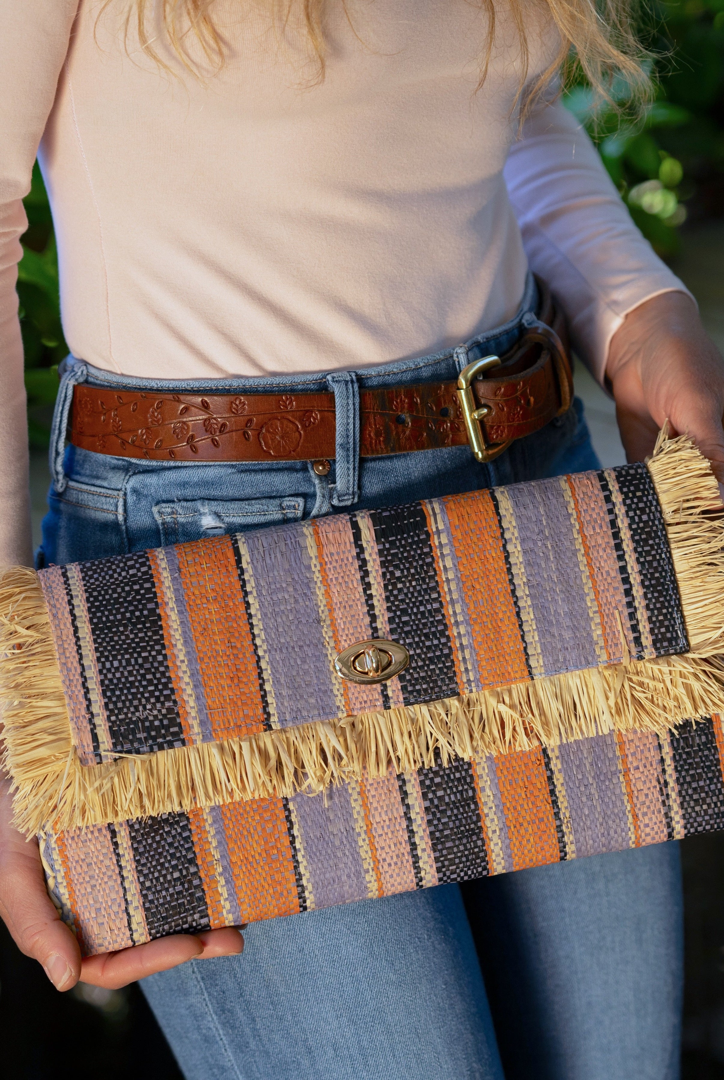 Model wearing Sophie Retro Stripe Multicolor Stripes Straw Clutch handmade loomed raffia purse handbag in muti-width vertical bands of denim retro/light/blue, dusk navy/dark/blue, light pink, coral orange/red and natural straw color with matching cross stitch edging and natural straw color raw fringe edge envelope shaped bag - Shebobo