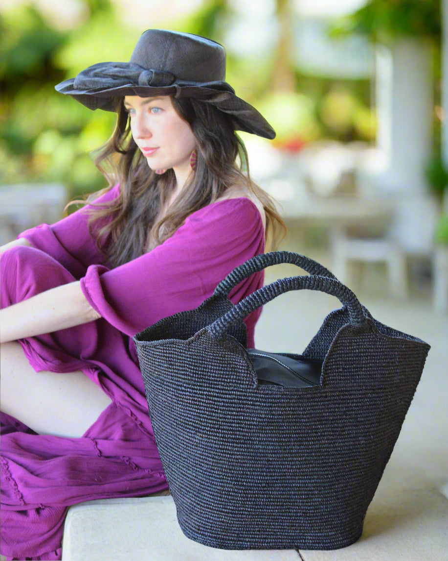 Model wearing Side view St. Tropez Heart Shaped Crochet Straw Basket Handbag handmade crochet natural raffia palm fiber in a solid hue of Black with a subtle link texture beach bag shoulder bag - Shebobo (with Lilly 5" Brim Fan Pleated Straw Sun Hats with Big Bow)