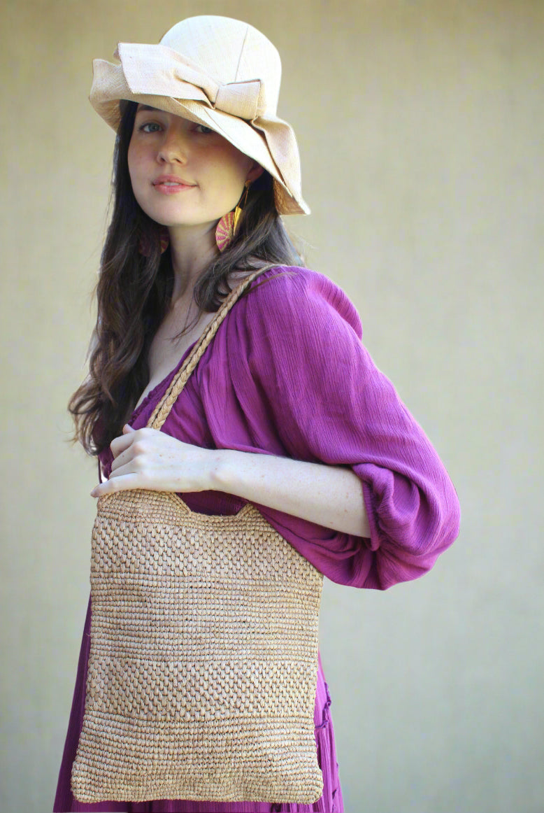Model wearing Tiana Crochet Straw Shoulder Bag Purse handmade crochet natural raffia palm fiber in a solid hue of Cappuccino tea/light/brown in subtle textured horizontal bands of two link patterns with matching braided handles - Shebobo (with Cara 3" Brim Bucket Straw Sun Hats with Big Bow)
