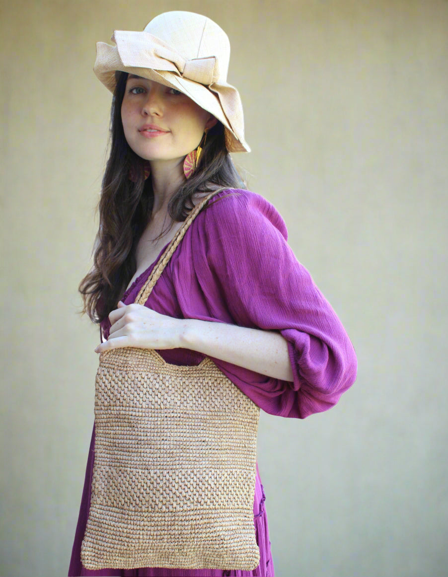 Model wearing Tiana Crochet Straw Shoulder Bag Purse handmade crochet natural raffia palm fiber in a solid hue of Cappuccino tea/light/brown in subtle textured horizontal bands of two link patterns with matching braided handles - Shebobo (with Cara 3" Brim Bucket Straw Sun Hats with Big Bow)