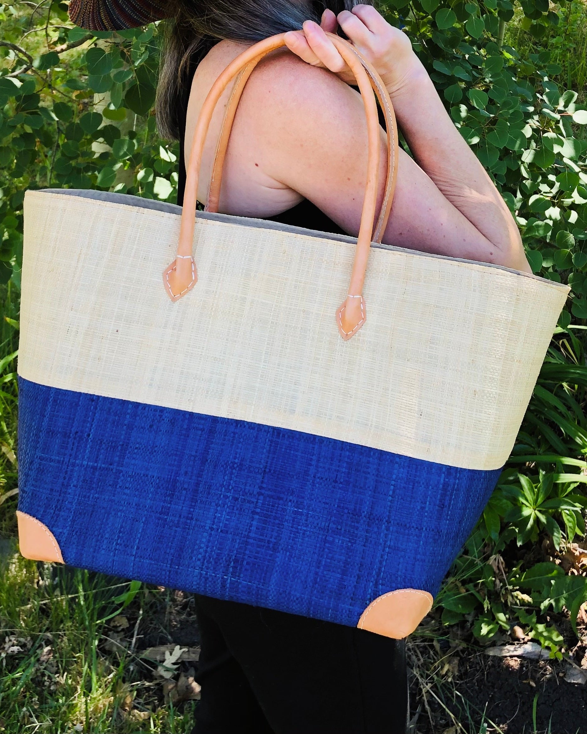 Model wearing Trinidad Natural and Azure Two Tone Straw Basket Bag handmade loomed raffia palm fiber in a two tone color block pattern with Azure dark/dusk/navy/blue on the bottom half and natural straw color on the top half of the bag with leather handles and accents beach bag tote purse - Shebobo