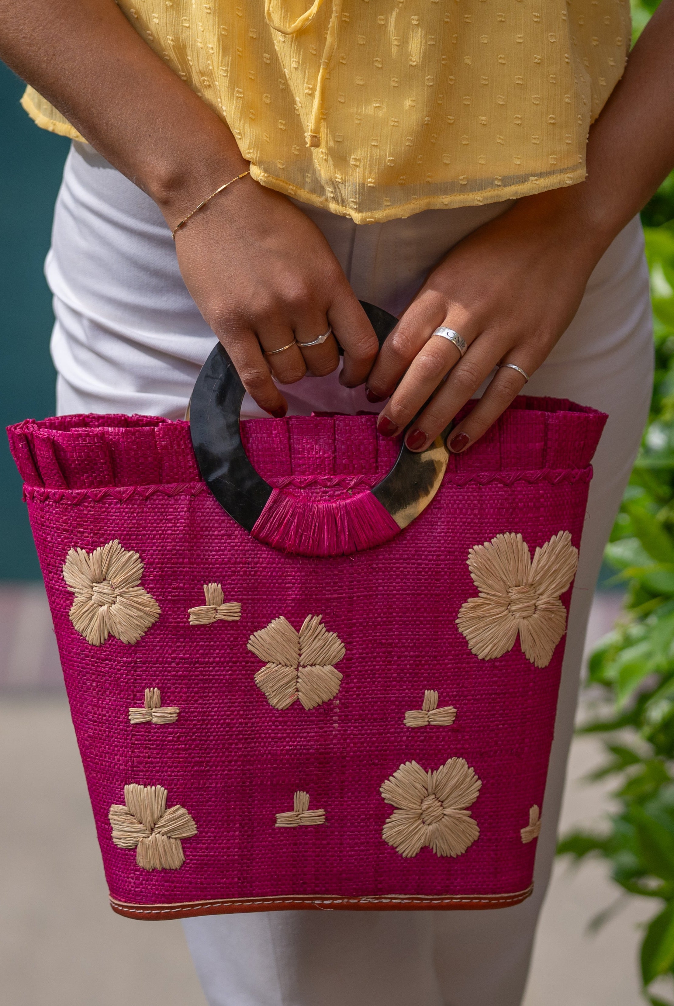 Model wearing Tulum Straw Bucket Bag with Flower Embroidery and Horn Handle Fuchsia Purse with Ruffle Edge handmade loomed raffia palm fiber in a solid hue of fuchsia hot/bright/barbie/pink with natural straw color hand embroidered floral pattern with multiple sized flowers and raffia ruffle on the opening handbag - Shebobo