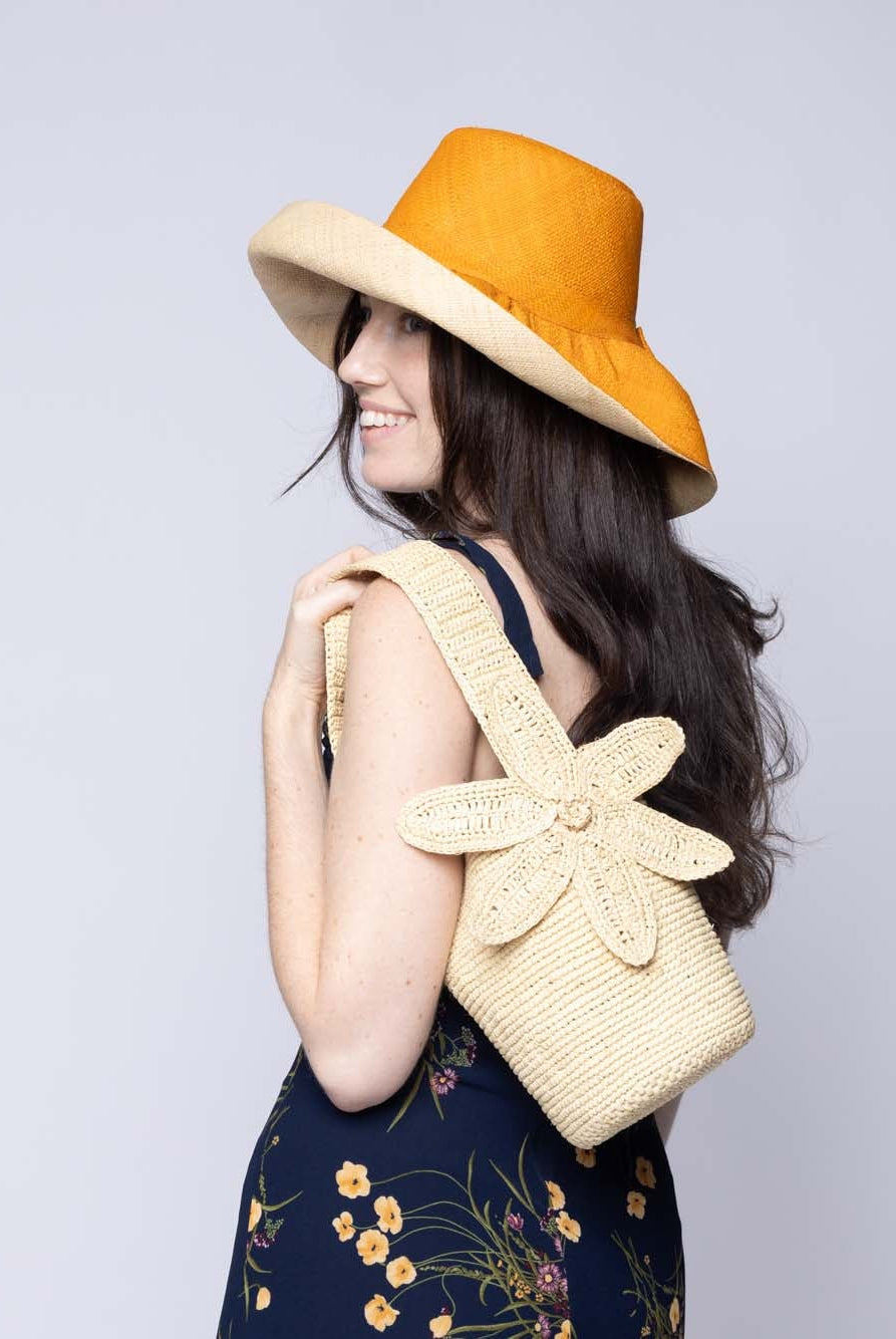 Model wearing Valencia Mini Crochet Petit Straw Basket Handbag with Flower Embellishment handmade natural raffia palm fiber in a solid hue of Natural straw color crochet in bands of horizontal link pattern texture with integrated strap handle and large flower embellishment - Shebobo
