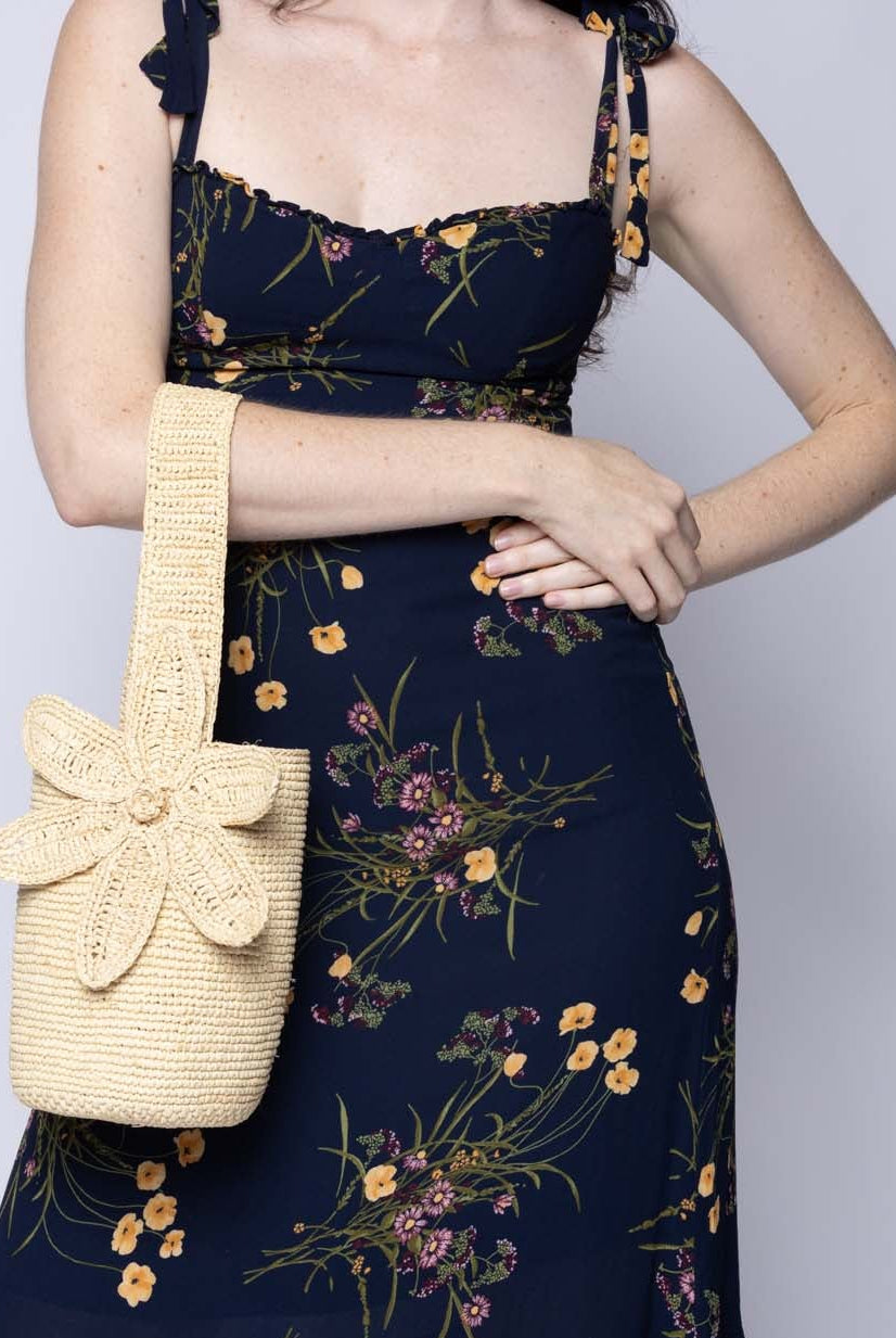 Model wearing Valencia Mini Crochet Petit Straw Basket Handbag with Flower Embellishment handmade natural raffia palm fiber in a solid hue of Natural straw color crochet in bands of horizontal link pattern texture with integrated strap handle and large flower embellishment - Shebobo