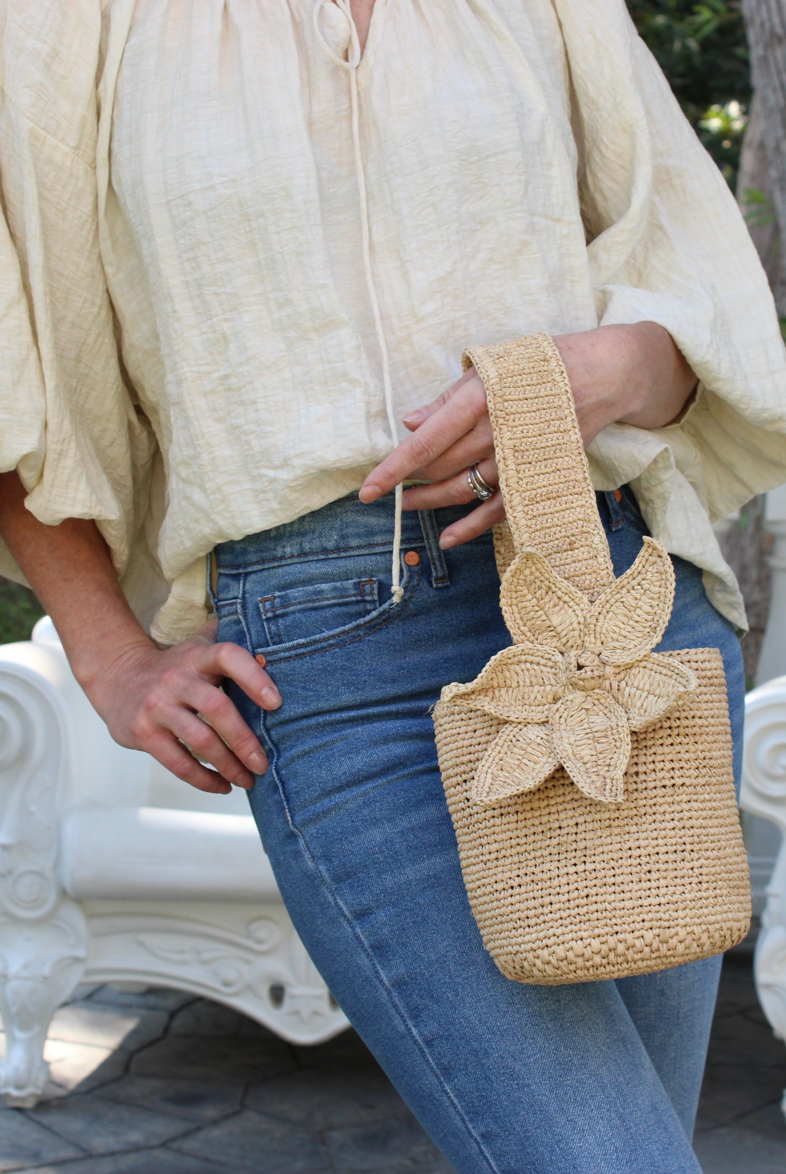 Model wearing Valencia Mini Crochet Petit Straw Basket Handbag with Flower Embellishment handmade natural raffia palm fiber in a solid hue of Natural straw color crochet in bands of horizontal link pattern texture with integrated strap handle and large flower embellishment - Shebobo