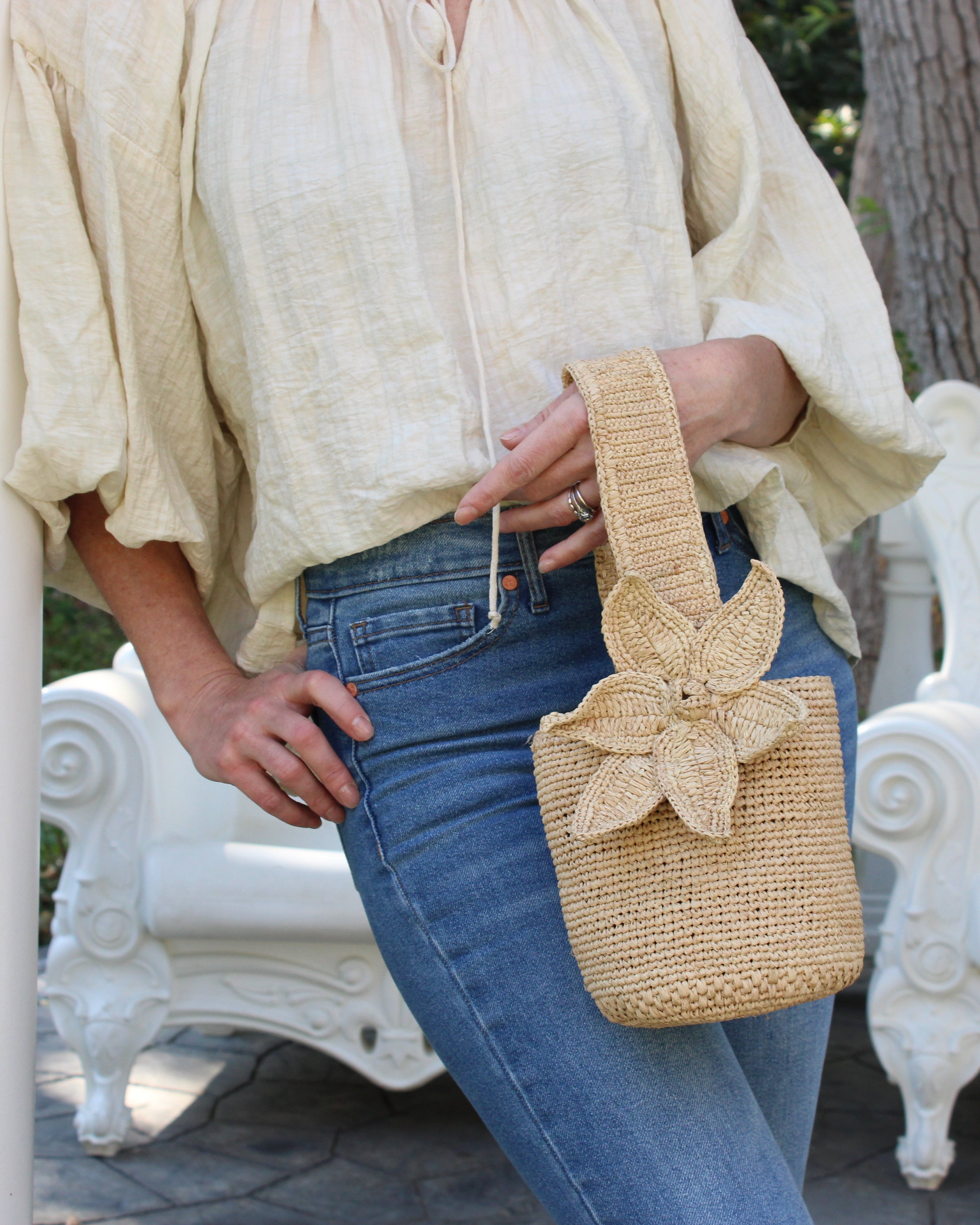 Model wearing Valencia Mini Crochet Petit Straw Basket Handbag with Flower Embellishment handmade natural raffia palm fiber in a solid hue of Natural straw color crochet in bands of horizontal link pattern texture with integrated strap handle and large flower embellishment - Shebobo