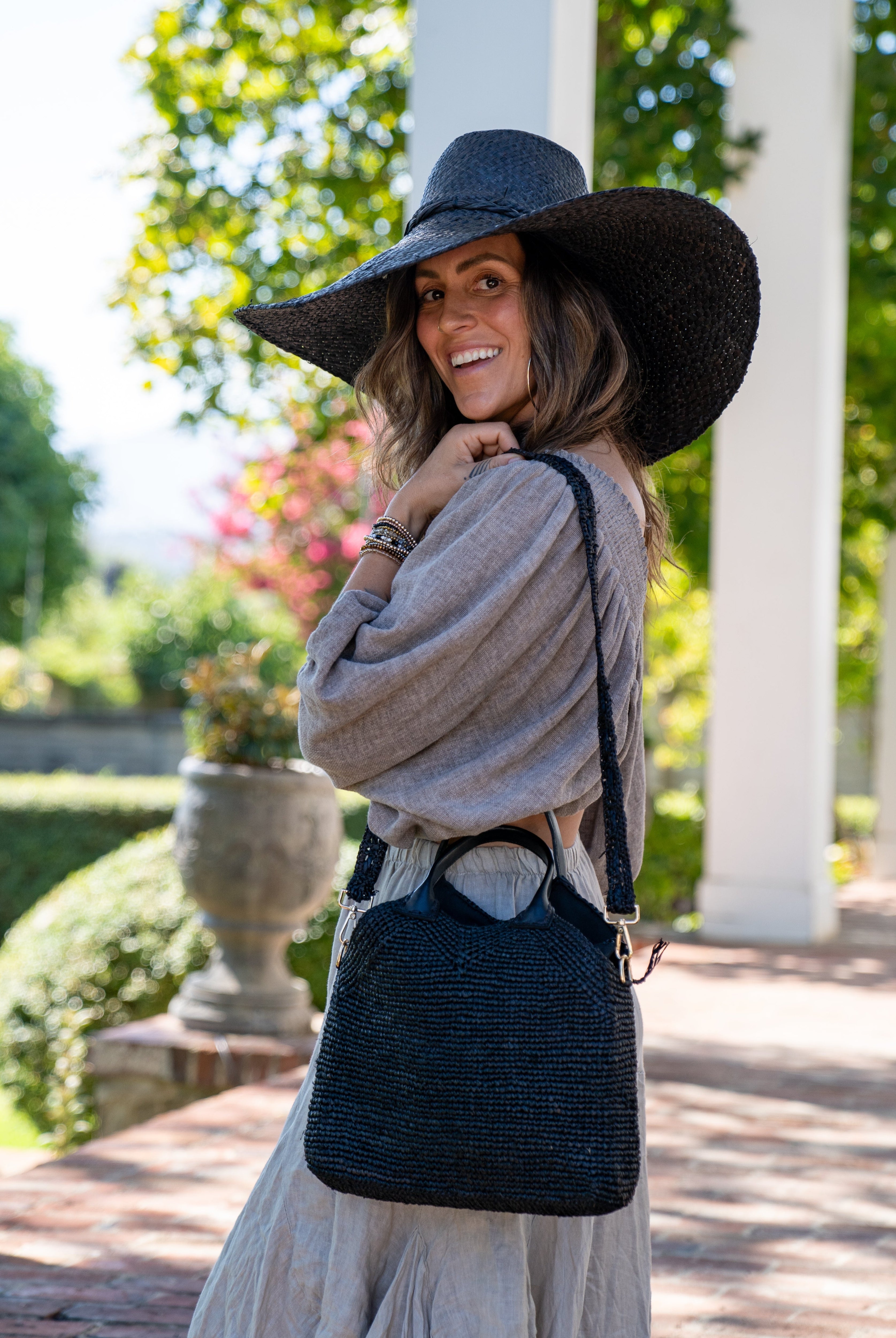 Model wearing Victoria Crochet Straw Handbag Convertible Crossbody Bag handmade crochet natural raffia palm fiber in a solid hue of black in a subtle texture of horizontal link bands pattern across the bag and m shape around the top of the bag with black leather handles and removable matching crossbody strap convertible purse - Shebobo (with Natasha 7" Wide Brim Woven XL Straw Sun Hats)