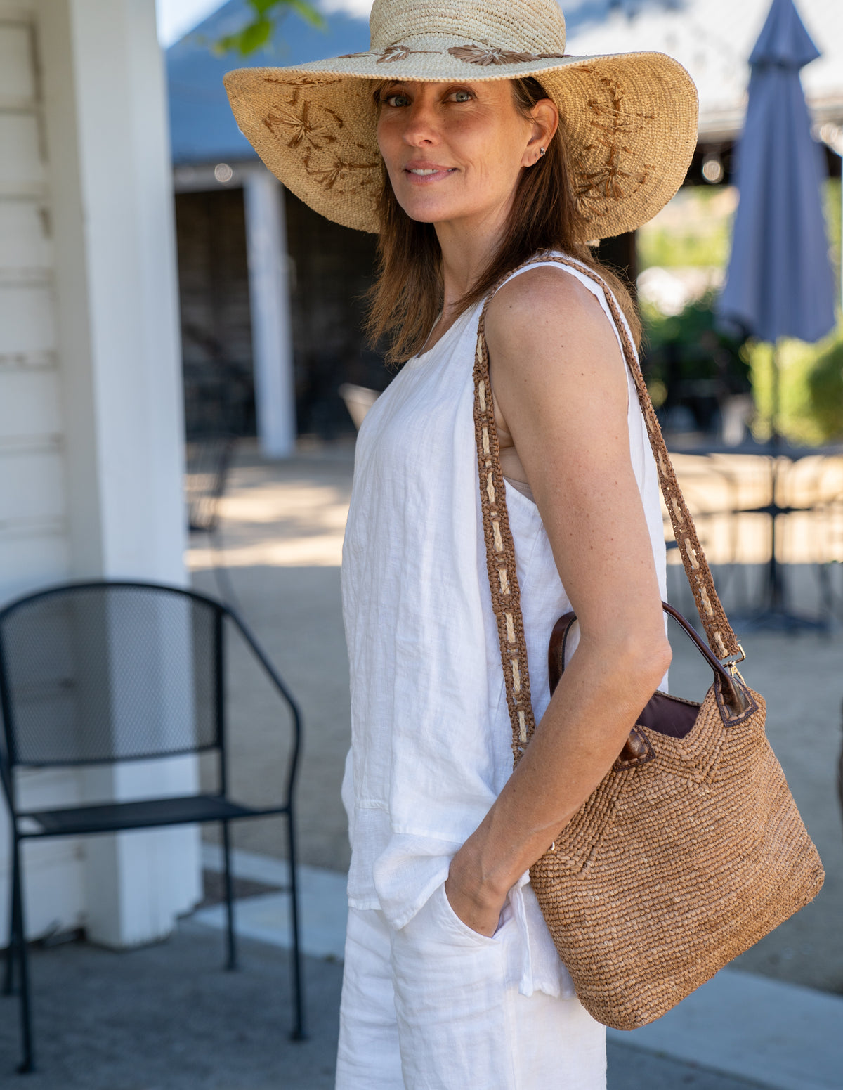 Model wearing Victoria Crochet Straw Handbag Convertible Crossbody Bag handmade crochet natural raffia palm fiber in a solid hue of cappuccino tea/light/brown in a subtle texture of horizontal link bands pattern across the bag and m shape around the top of the bag with dark brown leather handles and removable two tone natural and cappuccino tea/light/brown crossbody strap convertible purse - Shebobo (with Gigi 5.5" Brim Floral Embroidered Crochet Straw Sun Hats)