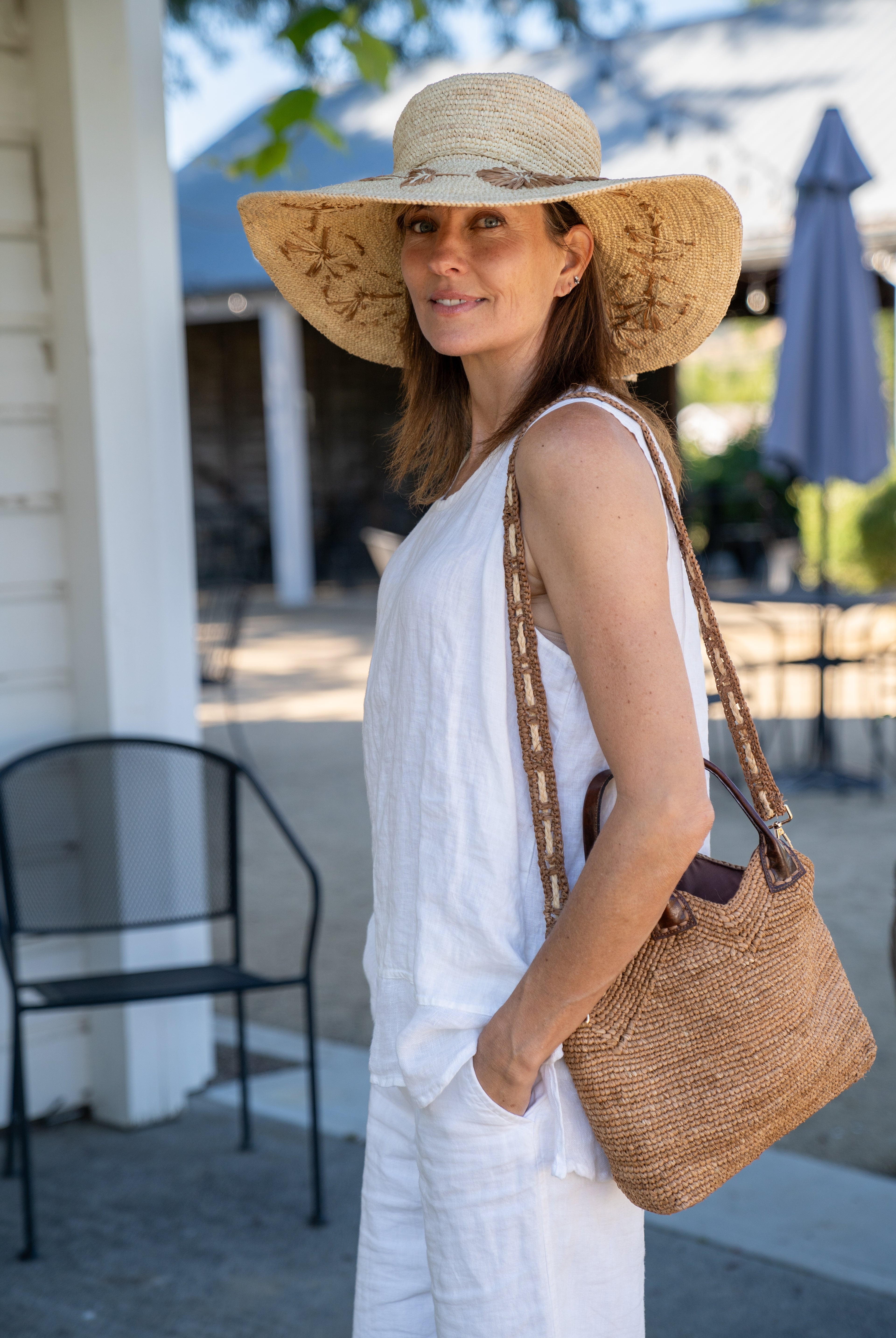 Model wearing Victoria Crochet Straw Handbag Convertible Crossbody Bag handmade crochet natural raffia palm fiber in a solid hue of cappuccino tea/light/brown in a subtle texture of horizontal link bands pattern across the bag and m shape around the top of the bag with dark brown leather handles and removable two tone natural and cappuccino tea/light/brown crossbody strap convertible purse - Shebobo (with Gigi 5.5" Brim Floral Embroidered Crochet Straw Sun Hats)