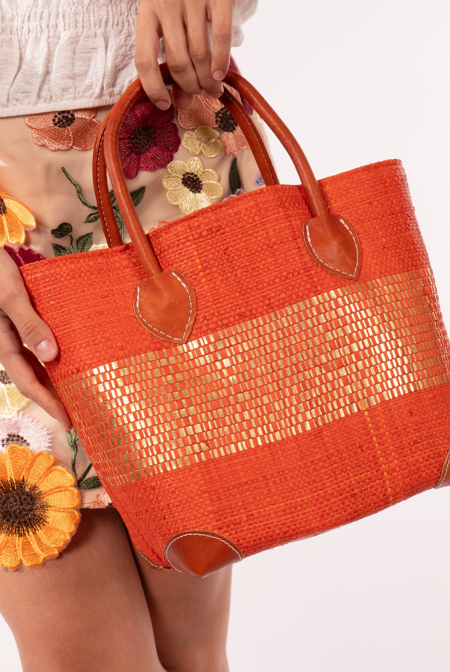Model wearing Wynwood Coral Straw Basket Bag with Metallic Detailing Handmade loomed raffia palm fiber in a solid hue of coral orange/red with a wide horizontal band of metallic strands woven in seamlessly handbag purse with leather handles - Shebobo