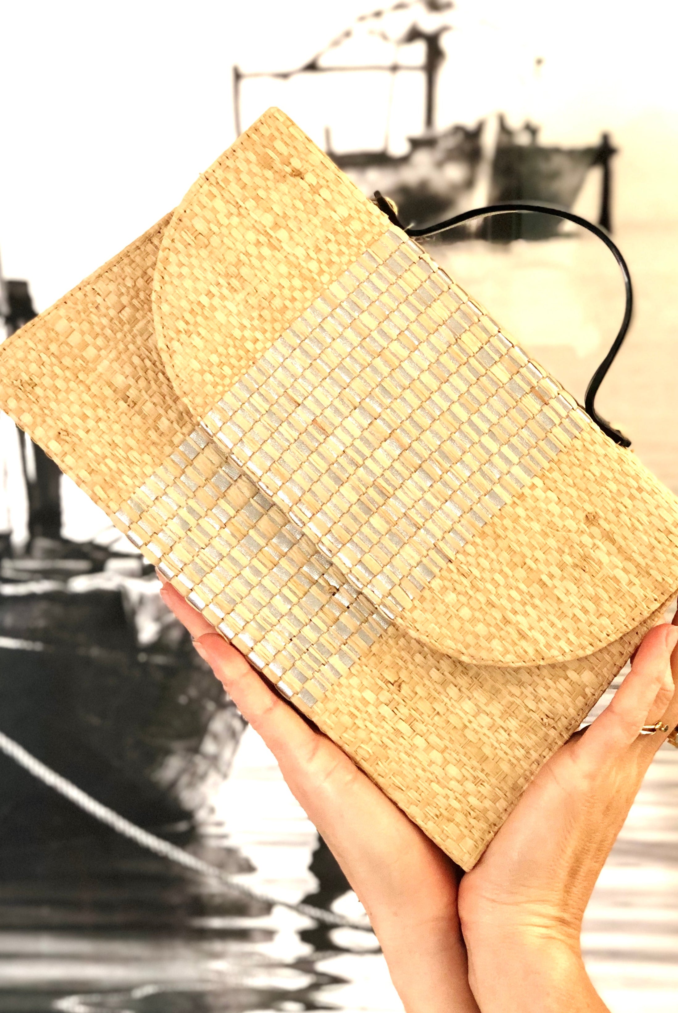 Model holding Wynwood Natural Straw Handbag with Metallic Detailing & Horn Handle handmade loomed raffia in natural straw color and silver metallic vegan leather in three vertical bands of color with the metallic weave centered on the purse under the handles - Shebobo