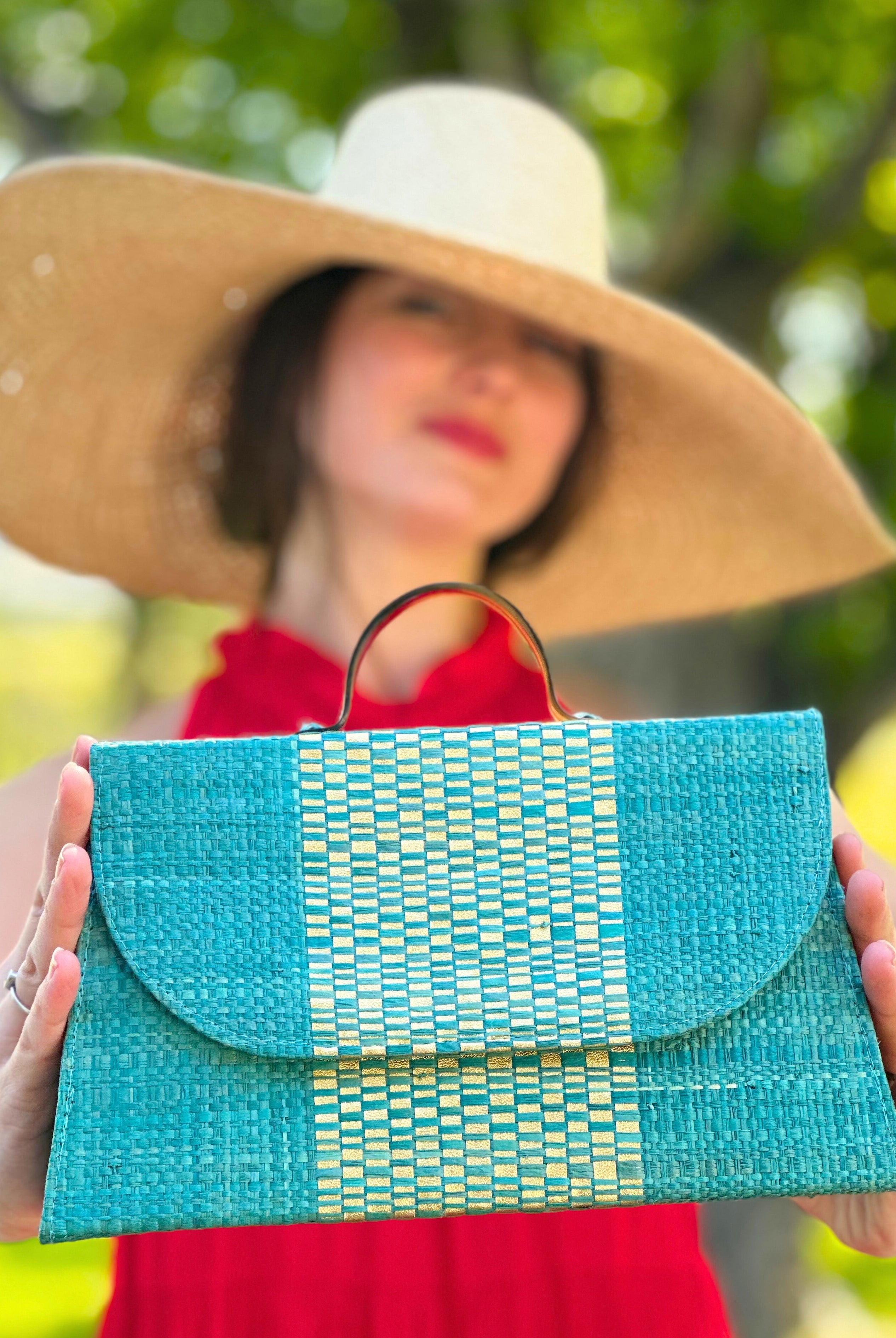 Model wearing Wynwood Turquoise Straw Handbag with Metallic Detailing & Horn Handle handmade loomed raffia in turquoise blue and metallic vegan leather in three vertical bands of color with the metallic weave centered on the purse under the handles with 7" Wide Brim Natasha Natural Woven Straw Sun Hat - Shebobo