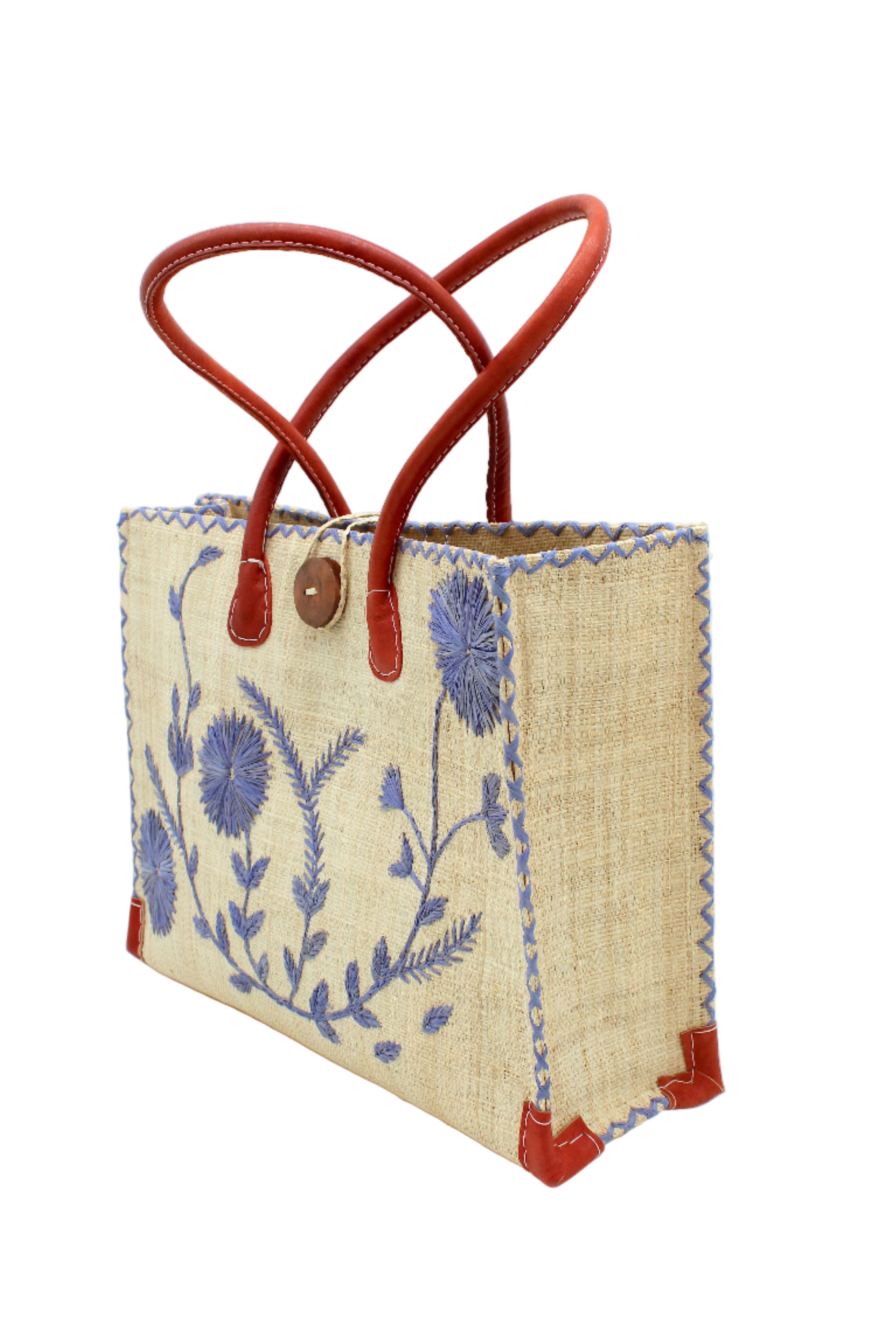 Side view Zafran Natural & Denim Embroidered Straw Beach Bag with Floral Pattern handmade natural raffia palm fiber in a solid hue of natural straw color with asymmetrical floral leaf, vine, and flower pattern in denim retro/light/blue with matching cross-stitch edging, leather handles, and wooden button closure shoulder bag handbag tote - Shebobo