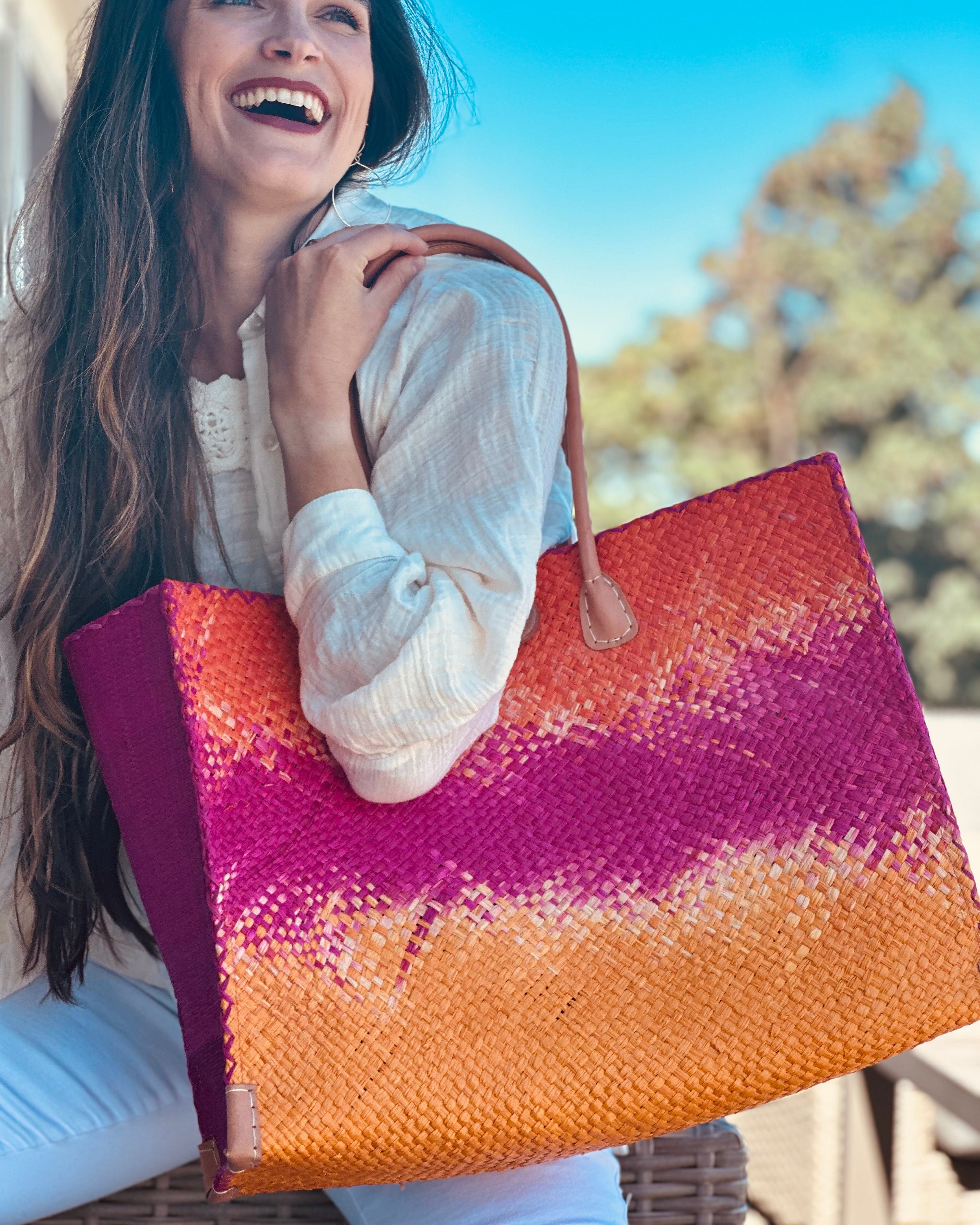 Model wearing Holden Fuchsia Ombre Zafran Straw Beach Bags handmade dip dyed raffia woven into a three part horizontal ombre pattern of coral orange/red on the top, fuchsia pink in the middle and both sides, and saffron yellow on the bottom with cross stitch edging, wooden button, and leather handles - Shebobo