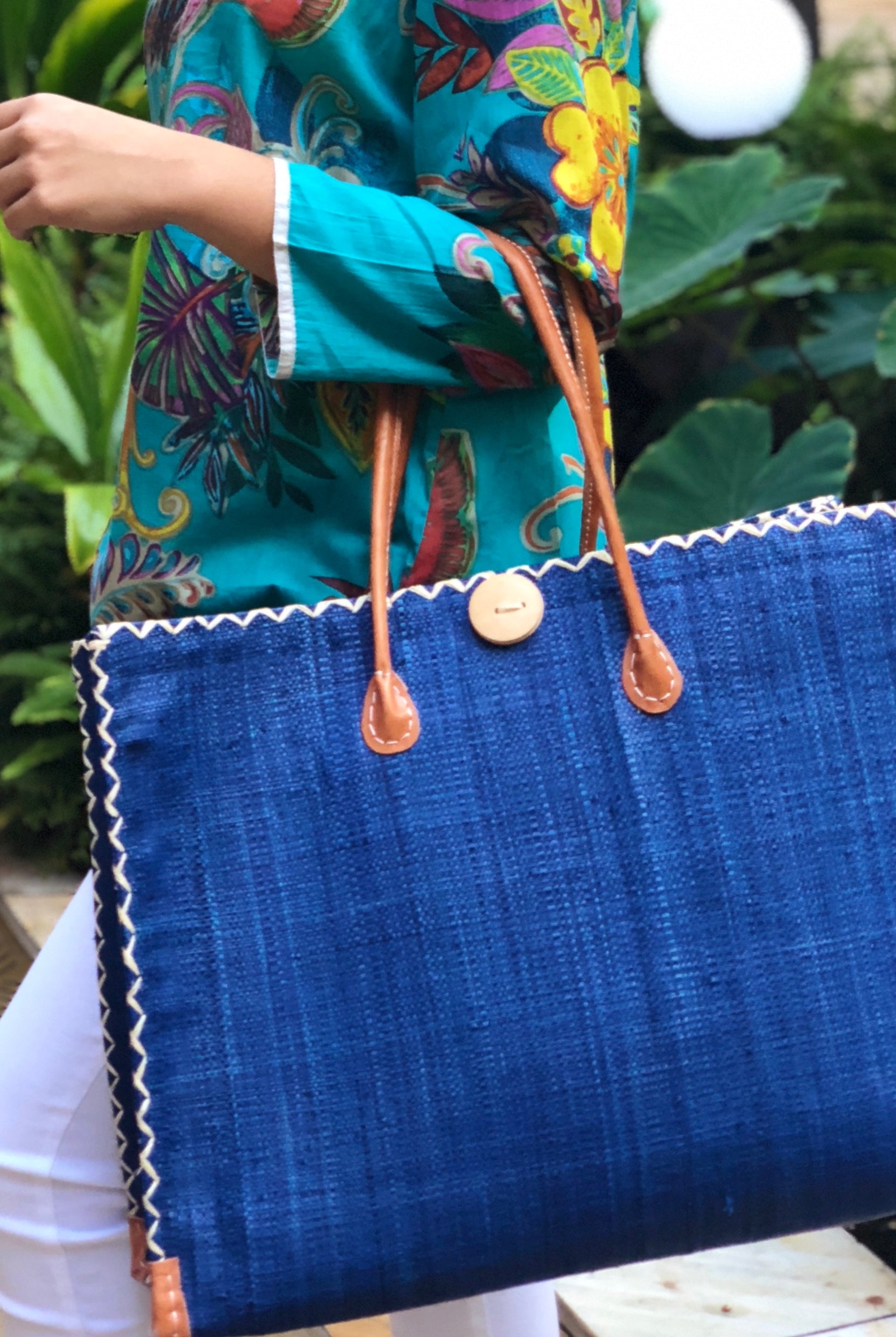 Model wearing Zafran Large Straw Beach Bag with Plastic Liner handmade loomed raffia in Navy deep blue with contrasting cross stitch edge binding, wood button closure, and leather handles & feet plus assorted print plastic lining - Shebobo