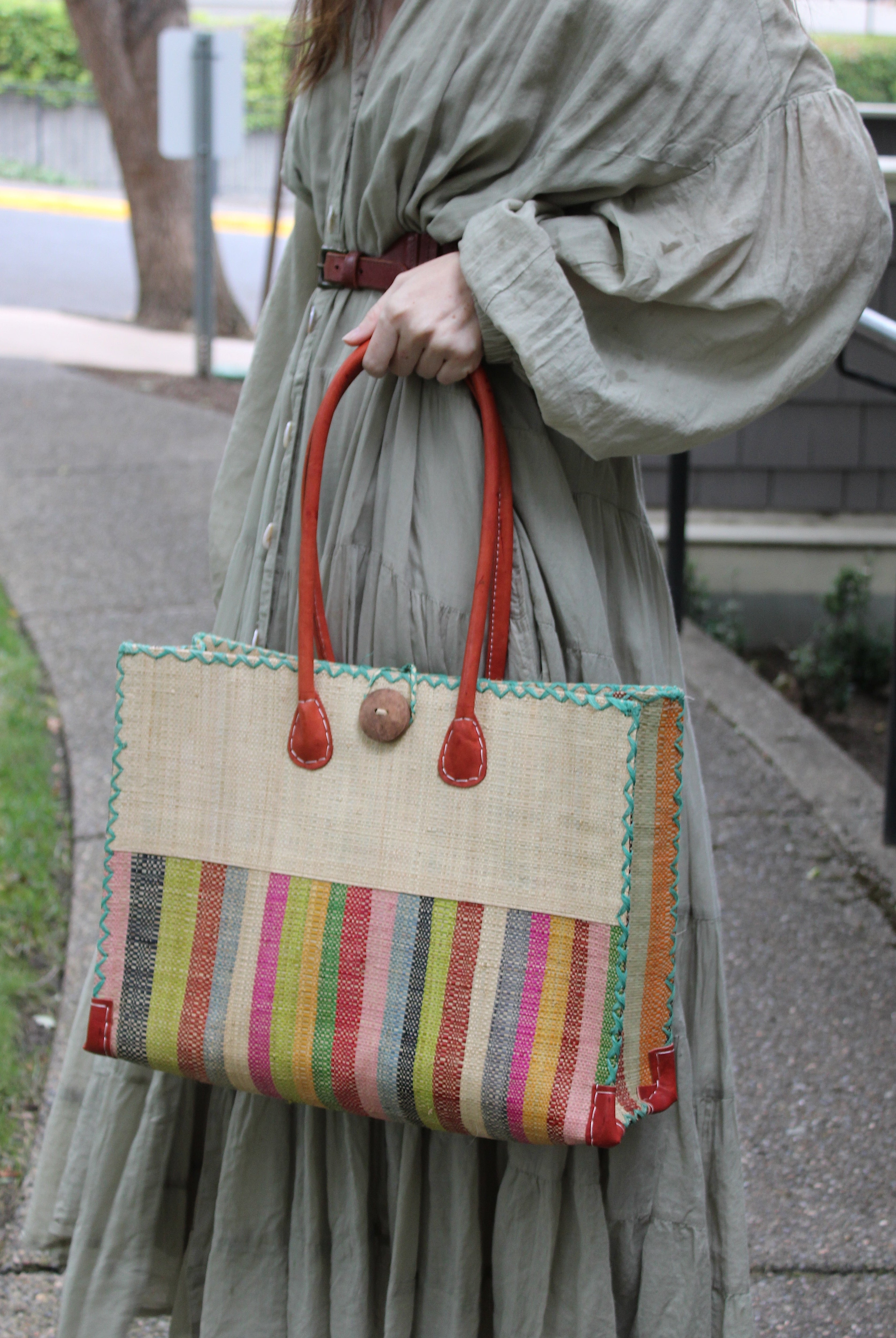 Model wearing Zafran Two Tone Wide Stripe Lollipop Stripe Straw Beach Bag handmade loomed raffia palm fiber in a solid hue of Natural straw color on the top half of the bag and a multicolor multi-width vertical stripe pattern of black, lime green, grass green, red, light/soft/baby pink, yellow, grey, whisky caramel/red/brown, light blue, fuchsia hot/bright/barbie/pink, and natural straw color on the bottom half of the bag with leather handles and feet and matching crosshatch stitch edging shoulder bag - She
