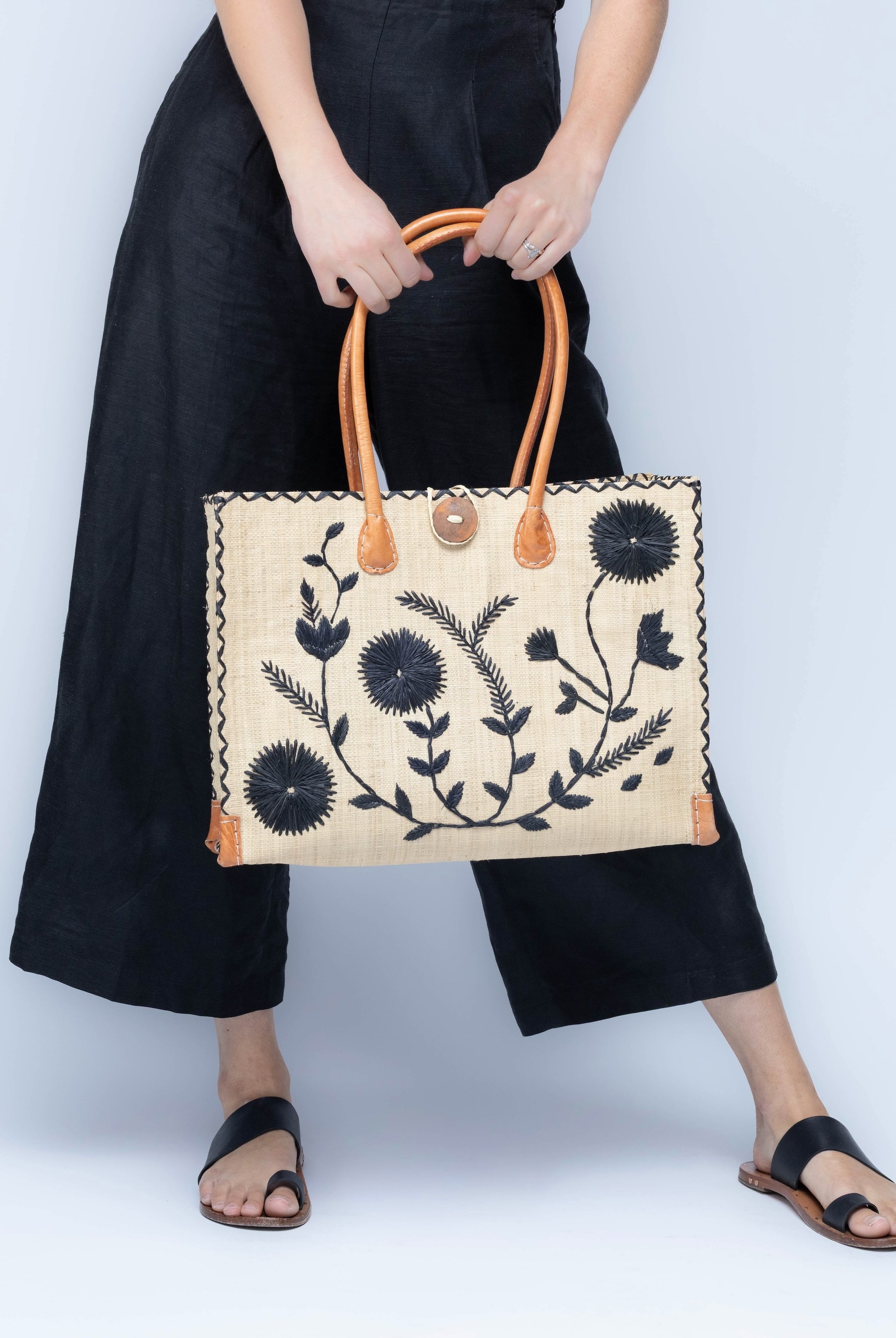Model wearing Zafran Natural & Black Embroidered Straw Beach Bag with Floral Pattern handmade natural raffia palm fiber in a solid hue of natural straw color with asymmetrical floral leaf, vine, and flower pattern in black with matching cross-stitch edging, leather handles, and wooden button closure shoulder bag handbag tote - Shebobo