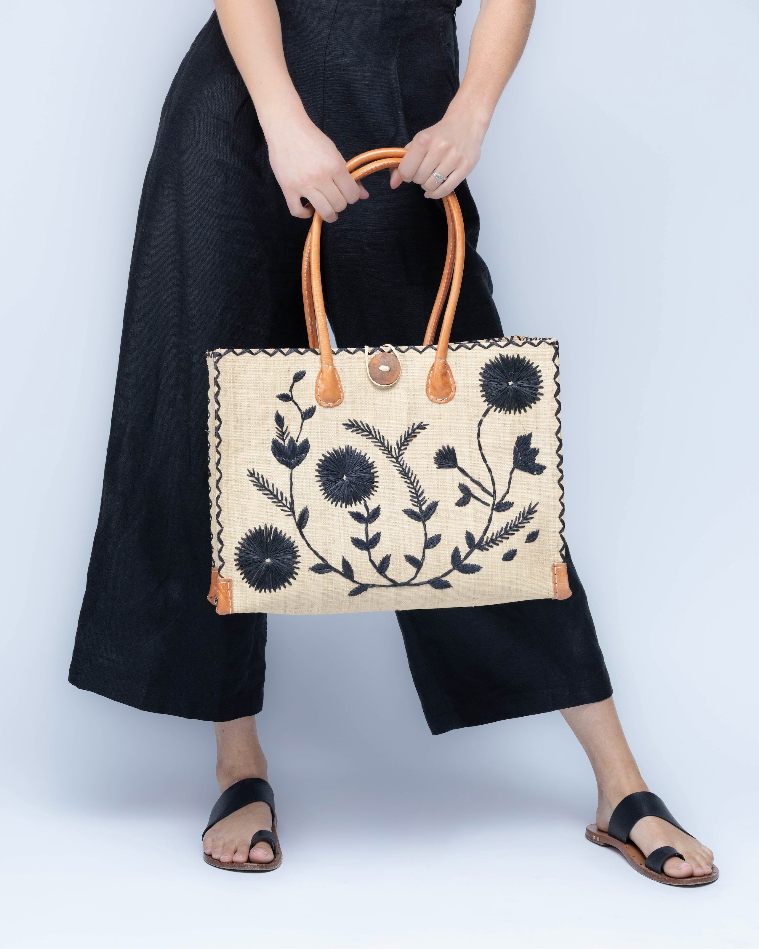 Model wearing Zafran Natural & Black Embroidered Straw Beach Bag with Floral Pattern handmade natural raffia palm fiber in a solid hue of natural straw color with asymmetrical floral leaf, vine, and flower pattern in black with matching cross-stitch edging, leather handles, and wooden button closure shoulder bag handbag tote - Shebobo