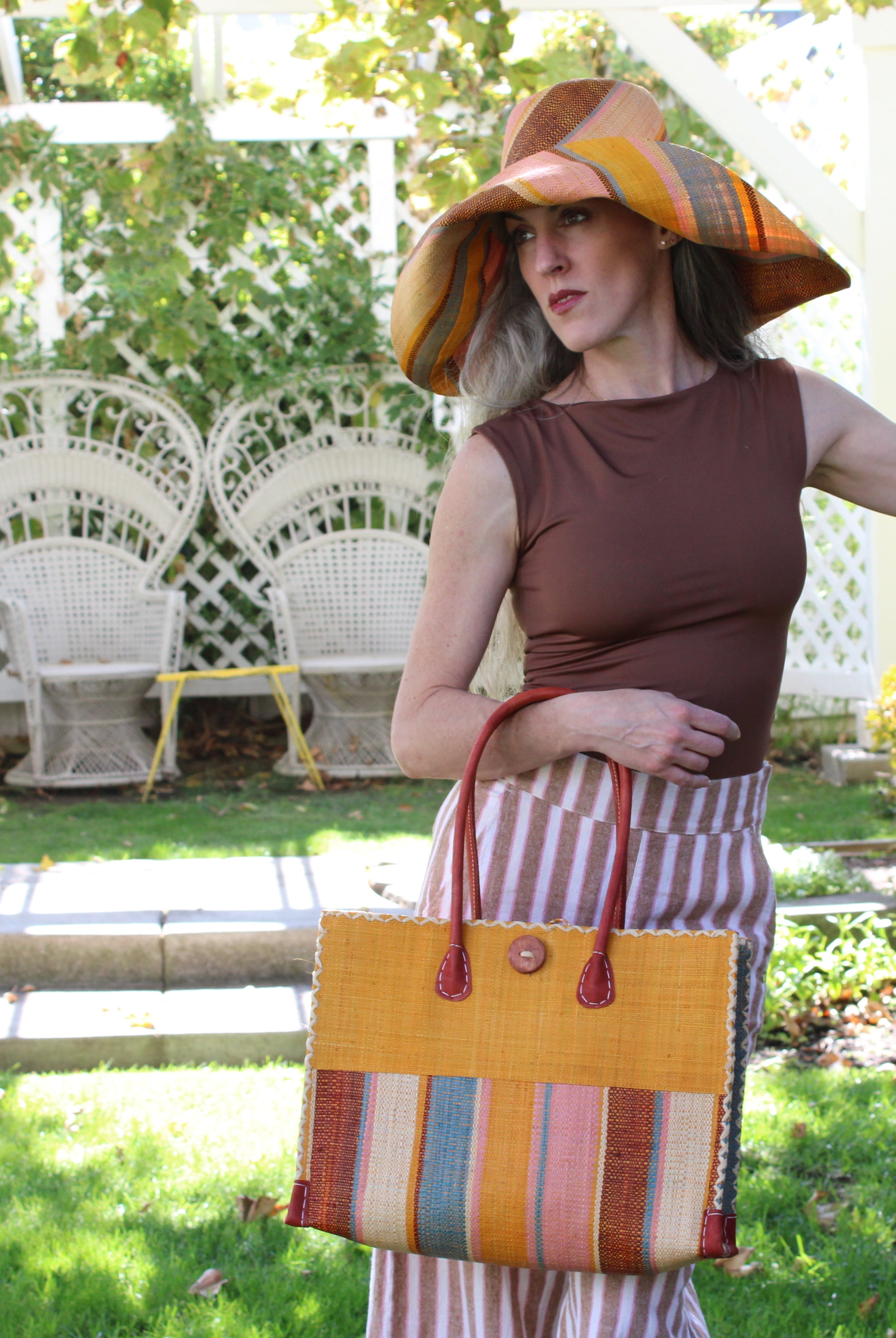 Model wearing Zafran Two Tone Wide Stripe Pink Blue Swirl Straw Beach Bag handmade loomed raffia palm fiber in a solid hue of Saffron yellow/orange on the top half of the bag and a multicolor multi-width vertical stripe pattern of black, soft/light/baby pink, light blue, saffron, and natural straw color on the bottom half of the bag with leather handles and feet and matching crosshatch stitch edging shoulder bag - Shebobo (with Monica 5" & 7" Wide Brim Multicolor Stripe Pattern Packable Straw Sun Hats)