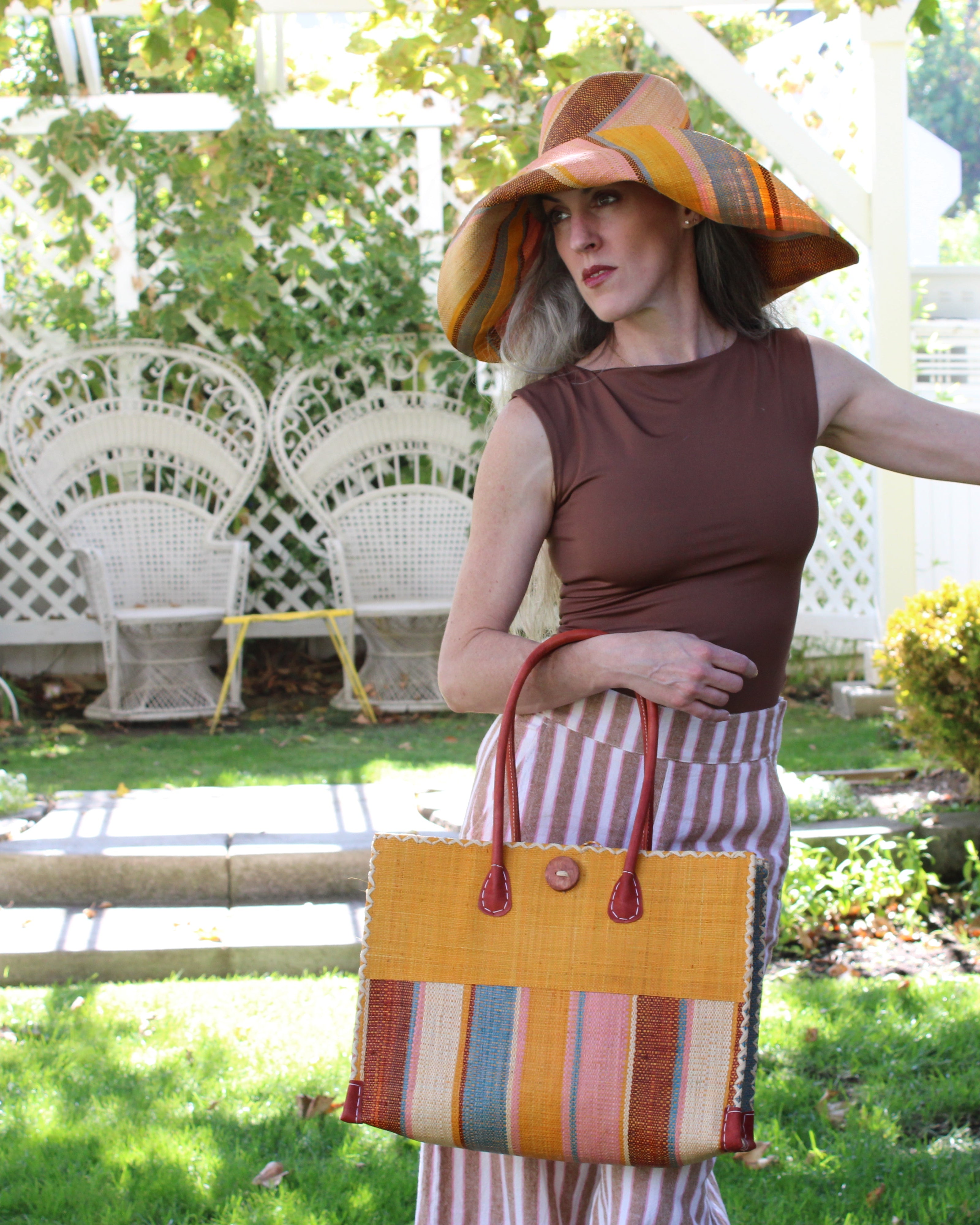 Model wearing Zafran Two Tone Wide Stripe Pink Blue Swirl Straw Beach Bag handmade loomed raffia palm fiber in a solid hue of Saffron yellow/orange on the top half of the bag and a multicolor multi-width vertical stripe pattern of black, soft/light/baby pink, light blue, saffron, and natural straw color on the bottom half of the bag with leather handles and feet and matching crosshatch stitch edging shoulder bag - Shebobo (with Monica 5" & 7" Wide Brim Multicolor Stripe Pattern Packable Straw Sun Hats)