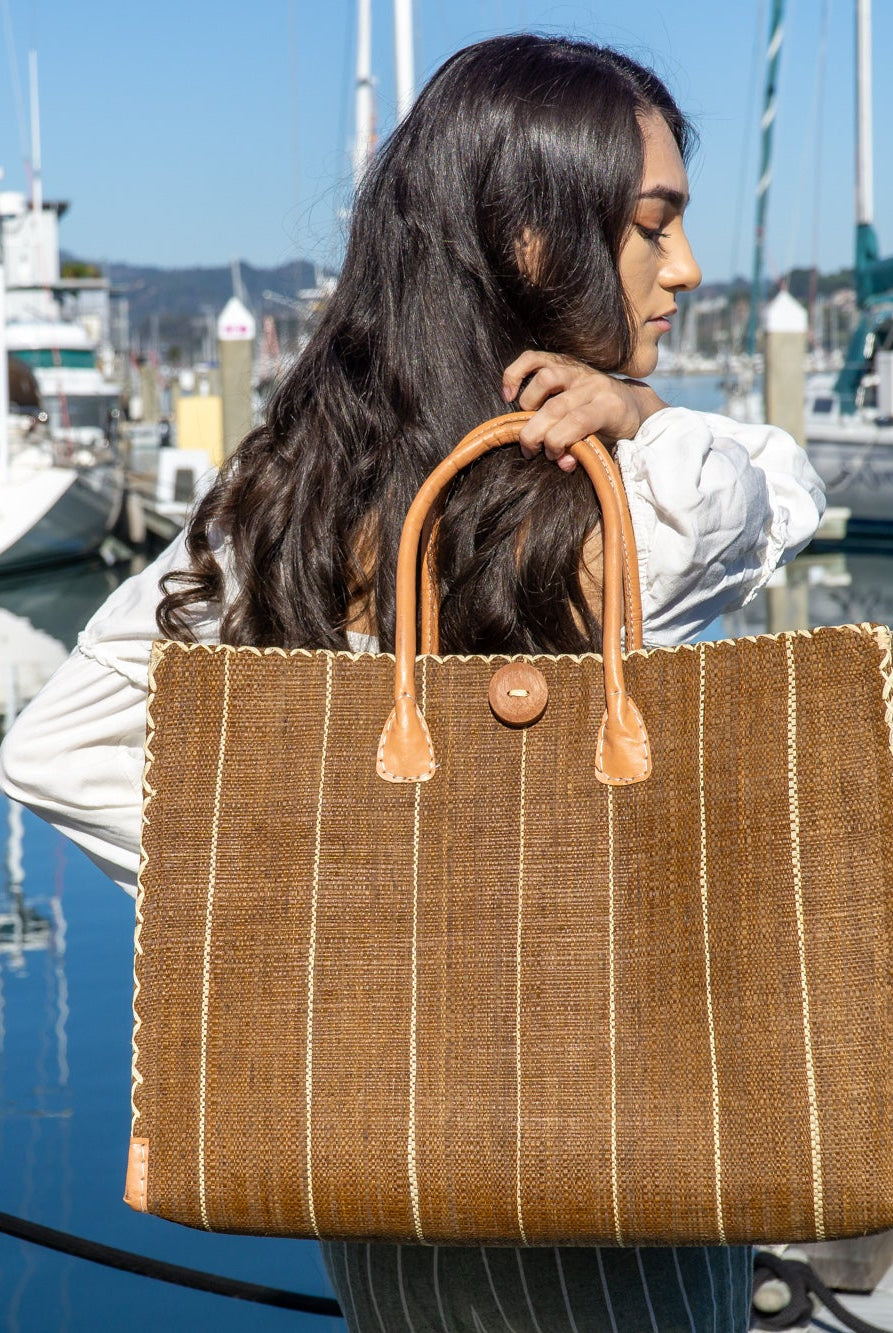 Model wearing Zafran Tobacco Pinstripes Straw Beach Bag with Plastic Liner handmade loomed raffia tote in wide bands of tobacco dark/cinnamon/brown with narrow bands of natural straw color that make a vertical pinstripe pattern with assorted print plastic lining and leather handles xl bag - Shebobo