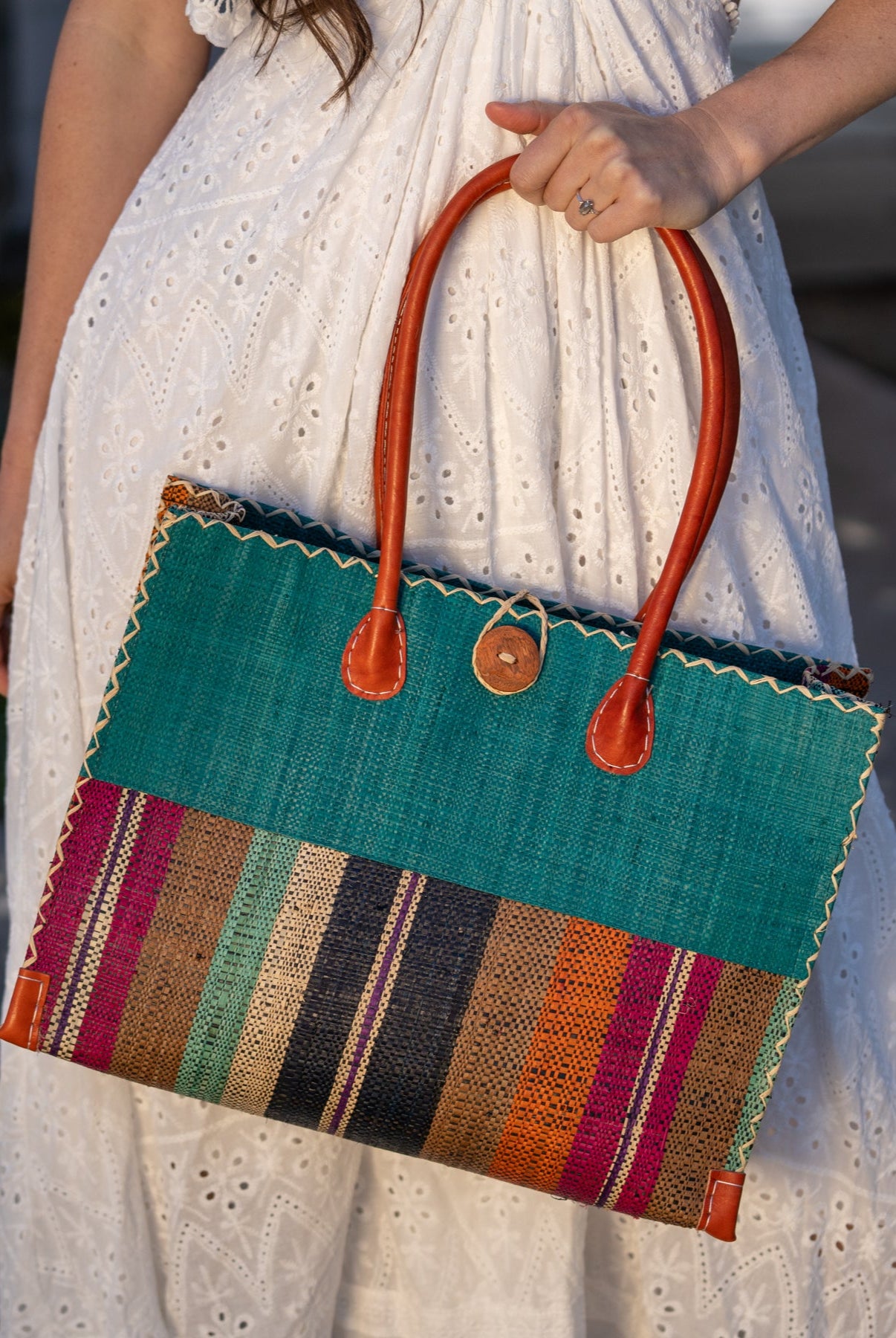 Model wearing Zafran Two Tone Wide Stripe Revel Swirl Straw Beach Bag handmade loomed raffia palm fiber in a solid hue of Teal dark/blue/green on the top half of the bag and a multicolor multi-width vertical stripe pattern of black, cinnamon tobacco/dark/brown, teal, coral orange/red, fuchsia hot/bright/barbie/pink, and natural straw color on the bottom half of the bag with leather handles and feet and matching crosshatch stitch edging shoulder bag - Shebobo