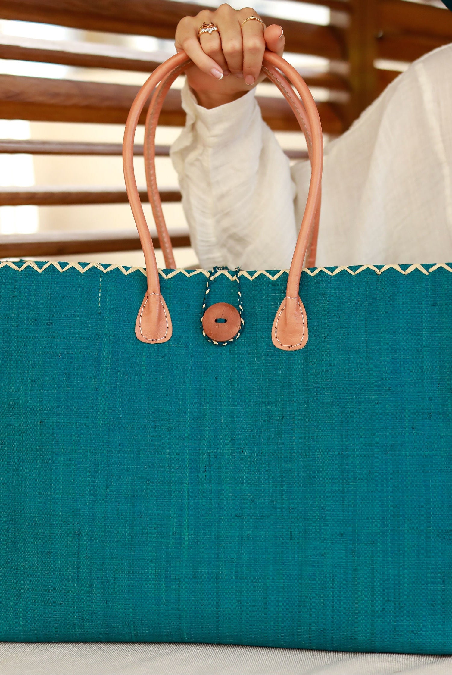 Model wearing Zafran Straw Beach Bag with Plastic Liner handmade loomed raffia in a solid hue of Turquoise blue/green with contrasting natural straw color cross stitch edge binding, wood button closure, and leather handles & feet plus assorted print plastic lining - Shebobo
