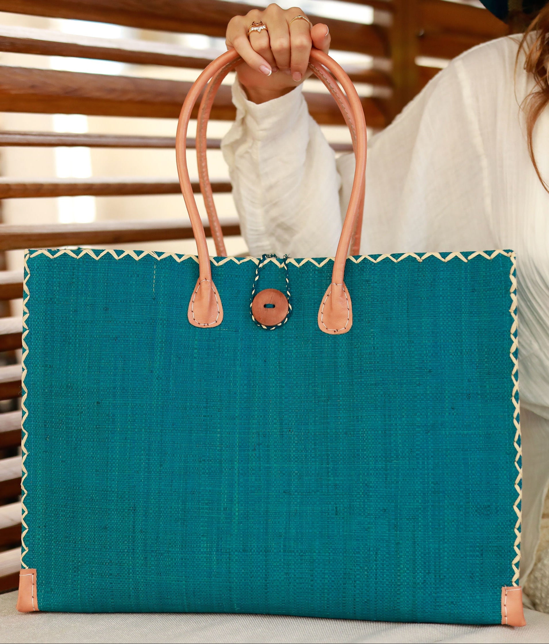 Model wearing Zafran Straw Beach Bag with Plastic Liner handmade loomed raffia in a solid hue of Turquoise blue/green with contrasting natural straw color cross stitch edge binding, wood button closure, and leather handles & feet plus assorted print plastic lining - Shebobo