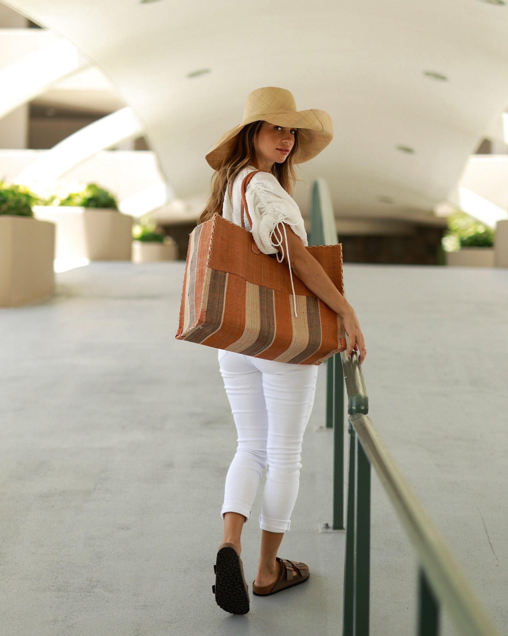 Model wearing Zafran Two Tone Wide Stripe Blush Swirl Straw Beach Bag handmade loomed raffia palm fiber in a solid hue of blush orange/pink on the top half of the bag and a multicolor multi-width vertical stripe pattern of blush, grey, and natural straw color on the bottom half of the bag with leather handles and feet and matching crosshatch stitch edging shoulder bag - Shebobo (with 7" Wide Brim Natural Packable Straw Sun Hat)