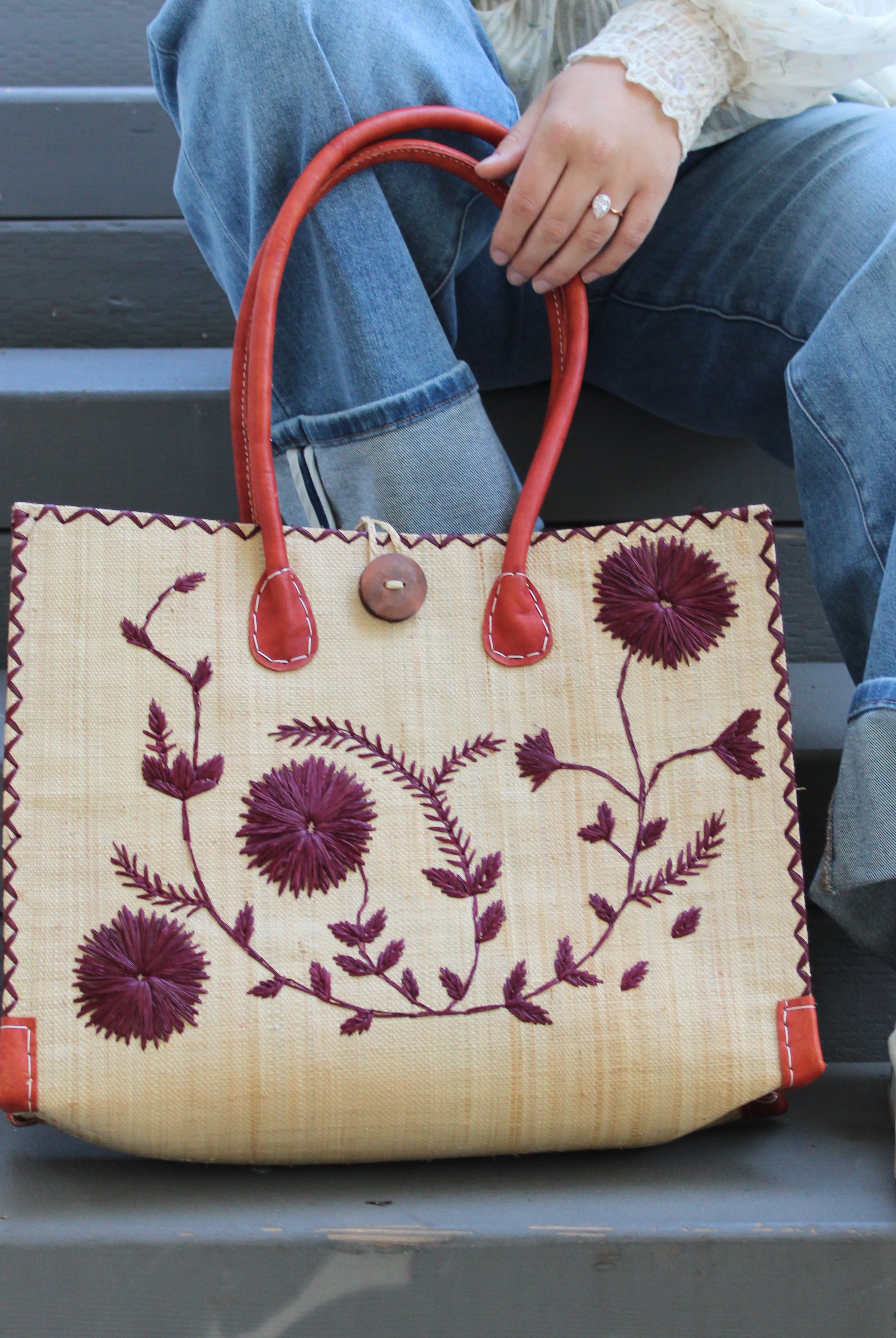 Model wearing Zafran Natural & Black Embroidered Straw Beach Bag with Floral Pattern handmade natural raffia palm fiber in a solid hue of natural straw color with asymmetrical floral leaf, vine, and flower pattern in umberwine burgundy/bordeaux/claret/dark/red/purple with matching cross-stitch edging, leather handles, and wooden button closure shoulder bag handbag tote - Shebobo