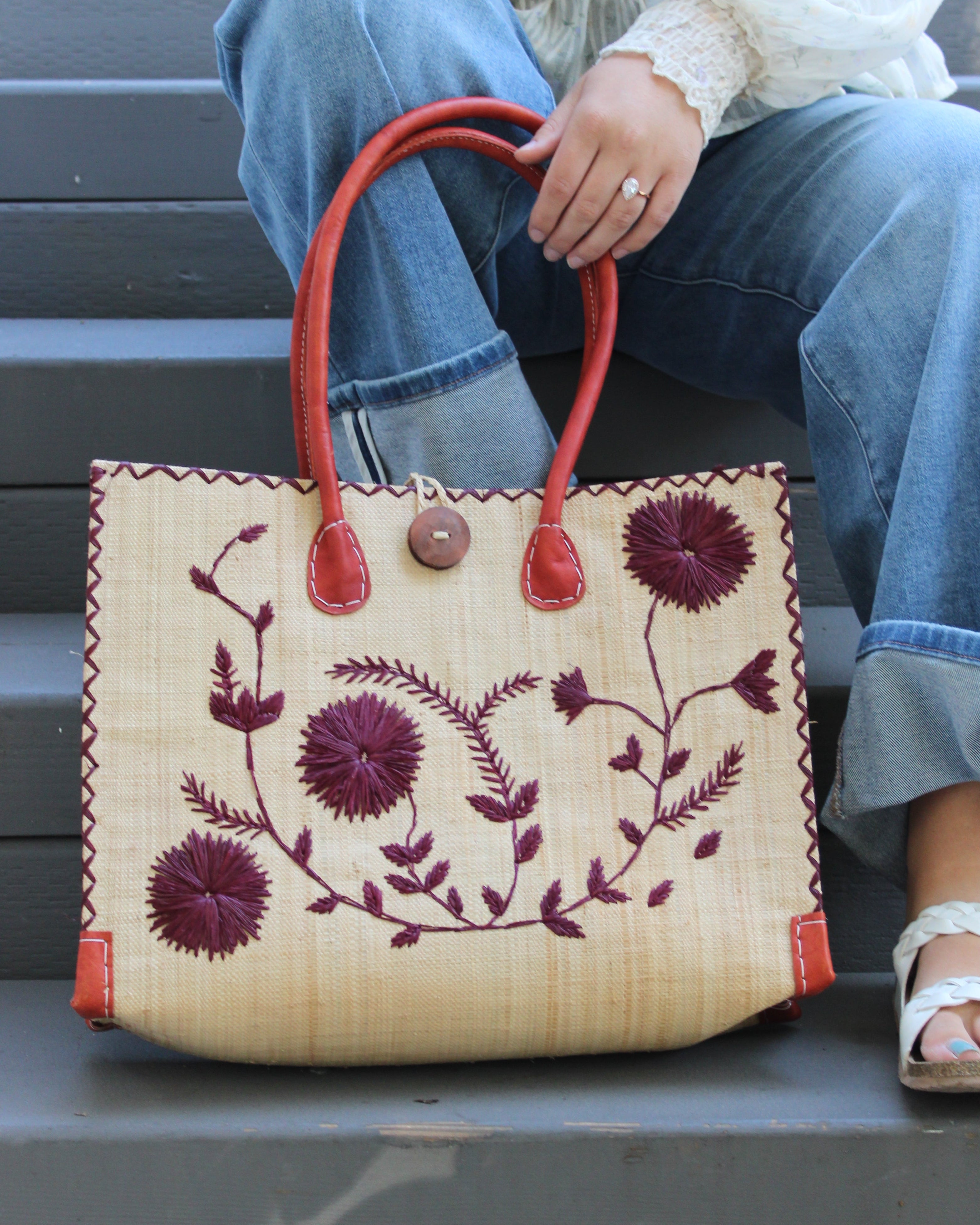 Model wearing Zafran Natural & Black Embroidered Straw Beach Bag with Floral Pattern handmade natural raffia palm fiber in a solid hue of natural straw color with asymmetrical floral leaf, vine, and flower pattern in umberwine burgundy/bordeaux/claret/dark/red/purple with matching cross-stitch edging, leather handles, and wooden button closure shoulder bag handbag tote - Shebobo