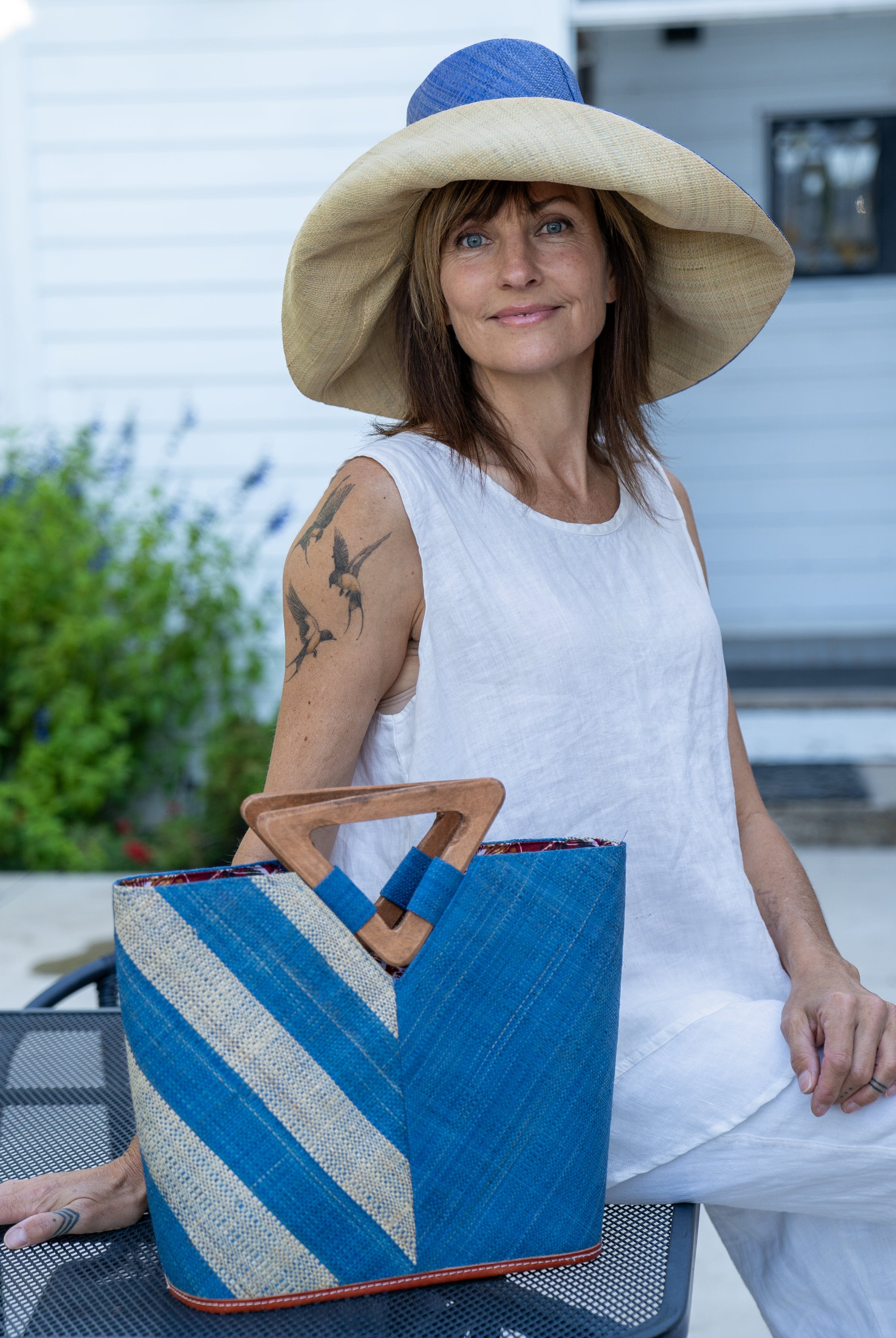 Model wearing Zuki Two Tone Denim Straw Handbag With Wood Triangle Handle handmade loomed raffia palm fiber in a two tone pattern with the left half of the bag even width stripes of denim retro/light/blue and natural straw color oriented as a diagonal stripe and the right half of the bag a solid hue of denim with carved wood handles - Shebobo (Monica 5" & 7" Wide Brim Two Tone Packable Straw Sun Hats)
