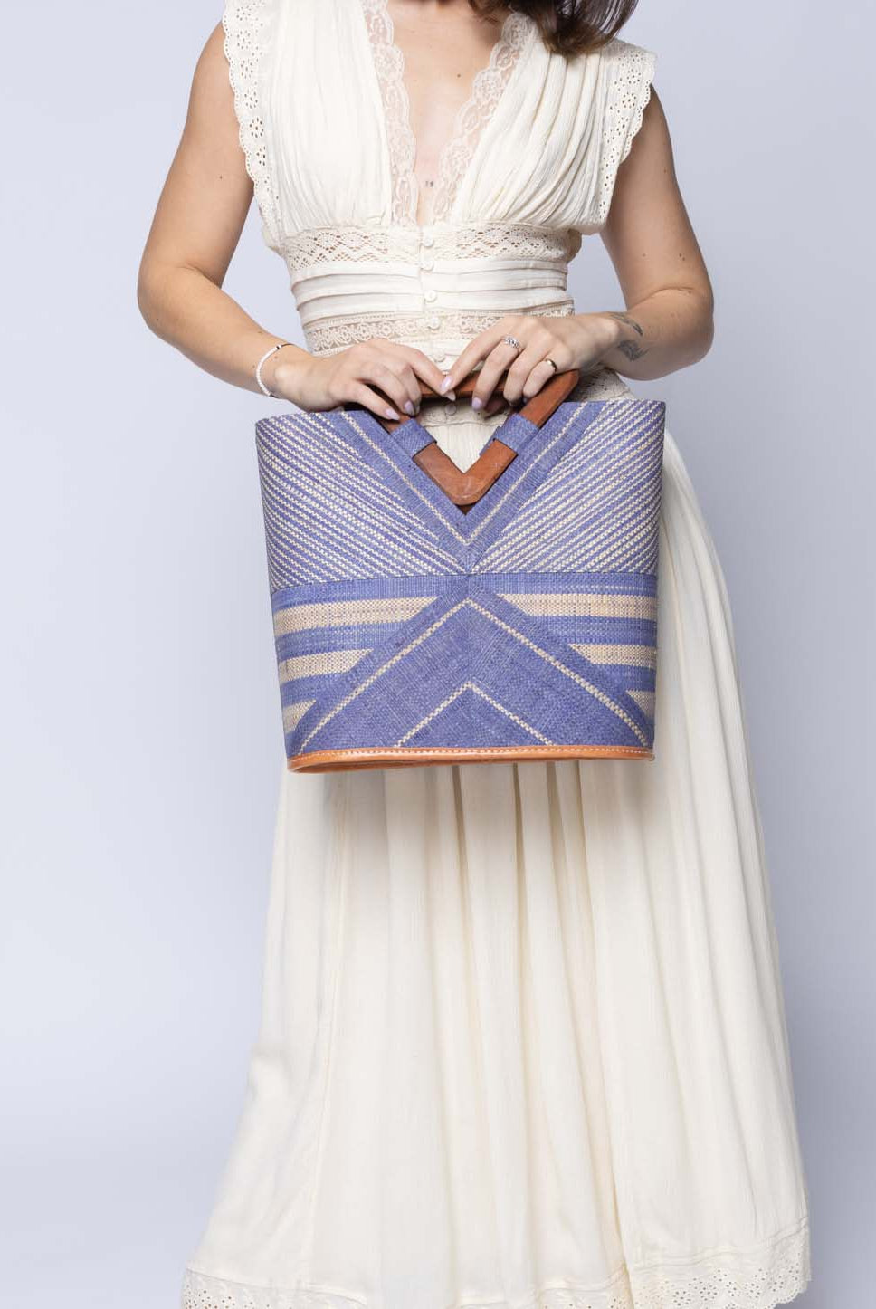 Model wearing Mariposa Zuki Raffia Straw Bucket Handbag with Wood Triangle Handles handmade loomed raffia palm fiber in a multi paneled pattern design of pinstripe, melange, and wide stripe combinations geometrically radiating from the center point of the bag outwards in a two tone color combination of Denim and Natural straw color with a solid hue of color on the back of the bag plus carved wooden handles purse - Shebobo