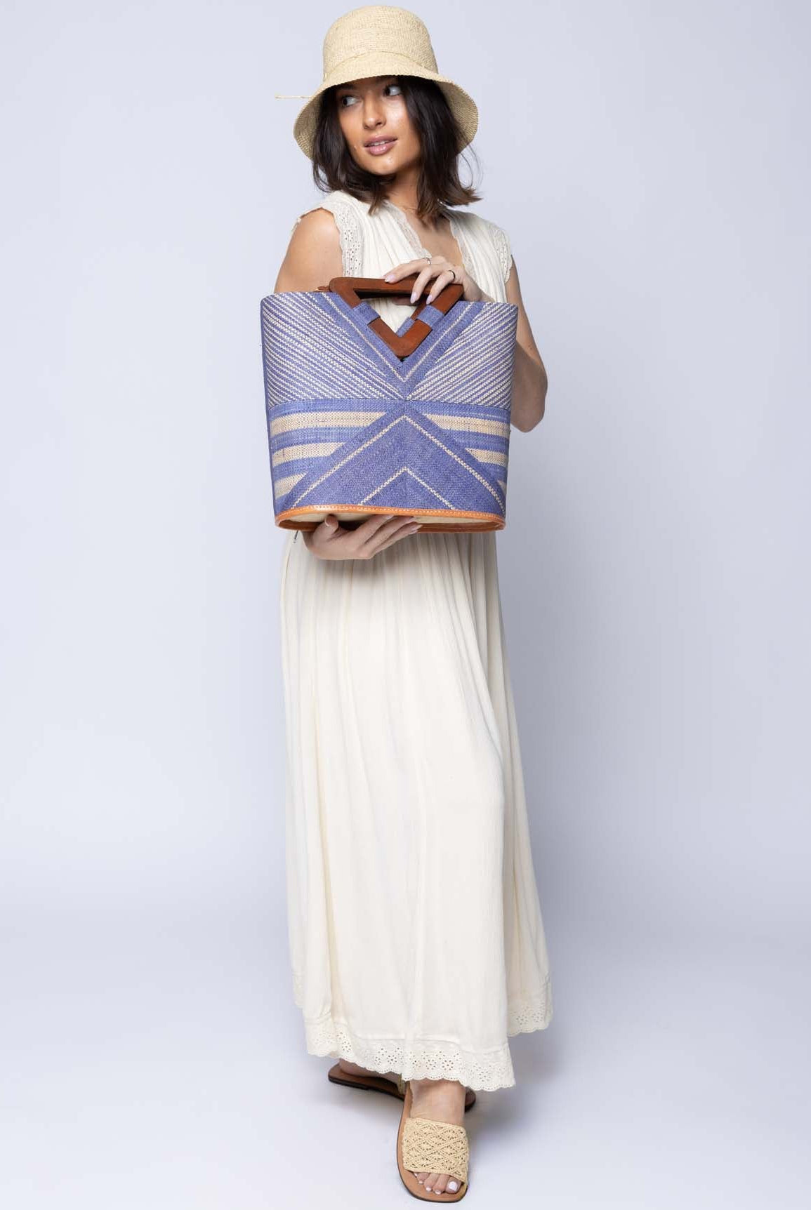 Model wearing Mariposa Zuki Raffia Straw Bucket Handbag with Wood Triangle Handles handmade loomed raffia palm fiber in a multi paneled pattern design of pinstripe, melange, and wide stripe combinations geometrically radiating from the center point of the bag outwards in a two tone color combination of Denim and Natural straw color with a solid hue of color on the back of the bag plus carved wooden handles purse - Shebobo