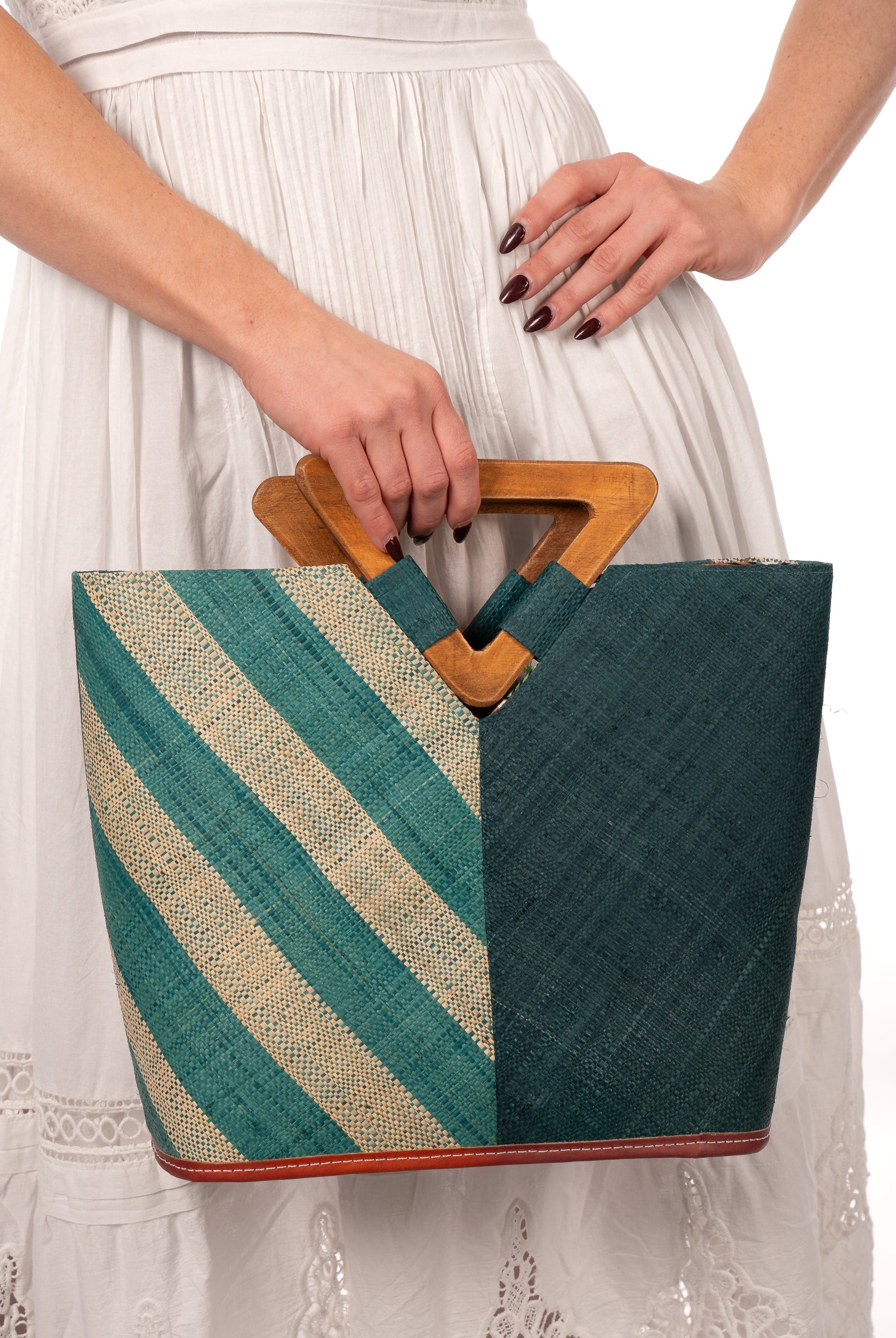 Model wearing Zuki Two Tone Teal Straw Handbag With Wood Triangle Handle handmade loomed raffia palm fiber in a two tone pattern with the left half of the bag even width stripes of teal dark/blue/green and natural straw color oriented as a diagonal stripe and the right half of the bag a solid hue of teal with carved wood handles - Shebobo