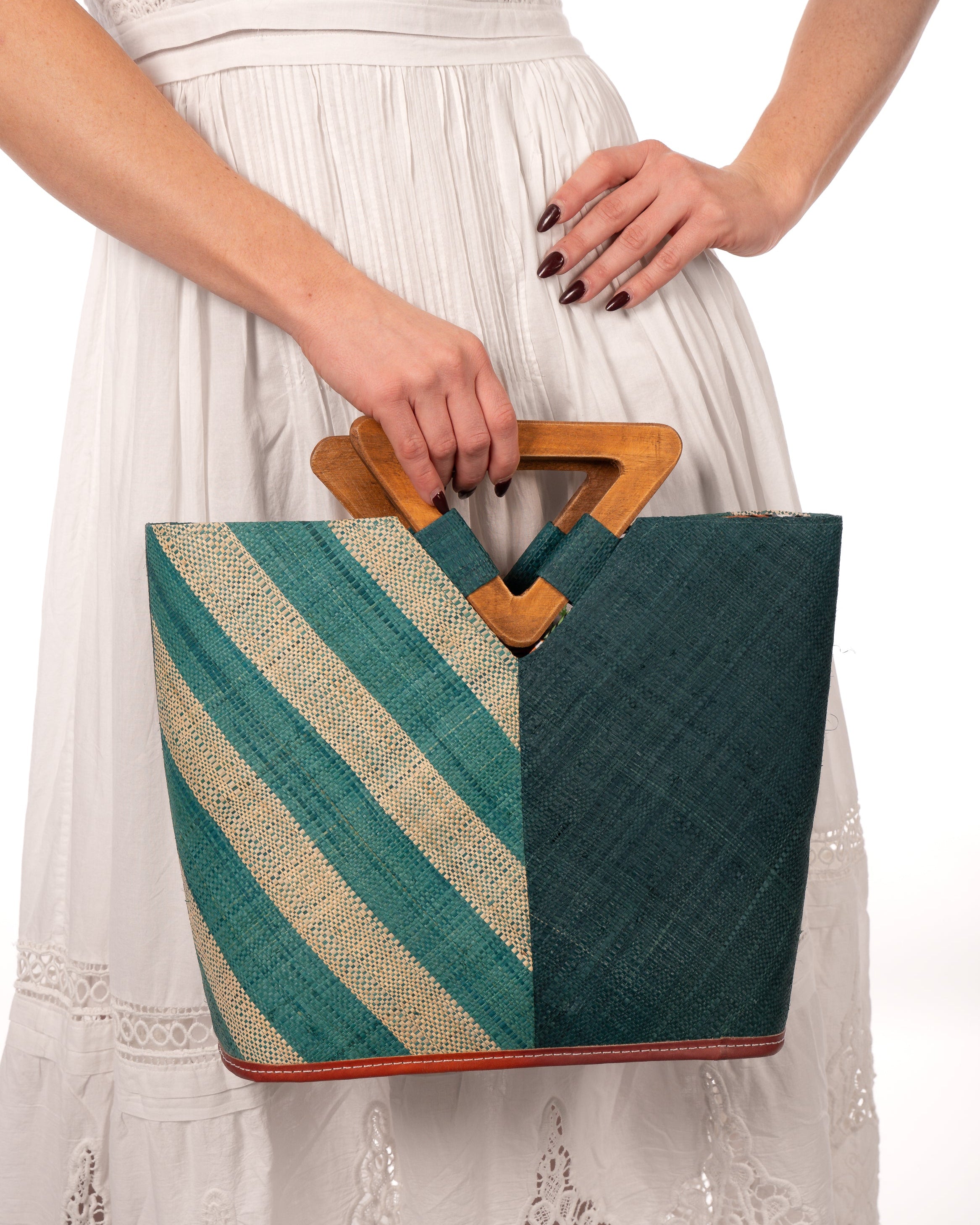 Model wearing Zuki Two Tone Teal Straw Handbag With Wood Triangle Handle handmade loomed raffia palm fiber in a two tone pattern with the left half of the bag even width stripes of teal dark/blue/green and natural straw color oriented as a diagonal stripe and the right half of the bag a solid hue of teal with carved wood handles - Shebobo