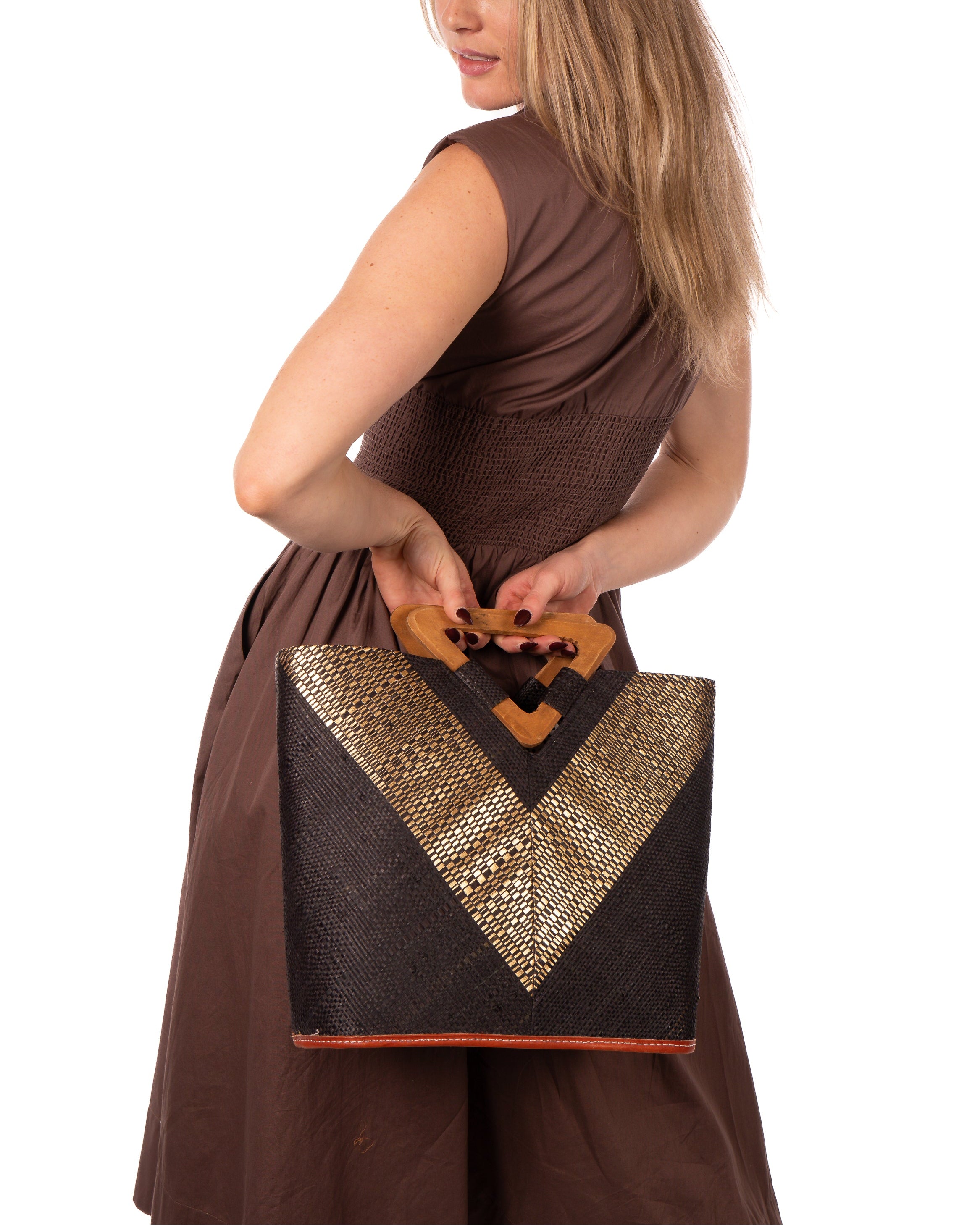 Model wearing Zuki Wynwood Black Straw Handbag with Metallic Detailing & Wood Triangle Handle handmade loomed raffia palm fiber in a solid hue of black with incorporated vegan leather gold metallic strands in a V pattern across the front and back of the bag with assorted print African wax cloth liners and carved wooden triangle handle purse - Shebobo