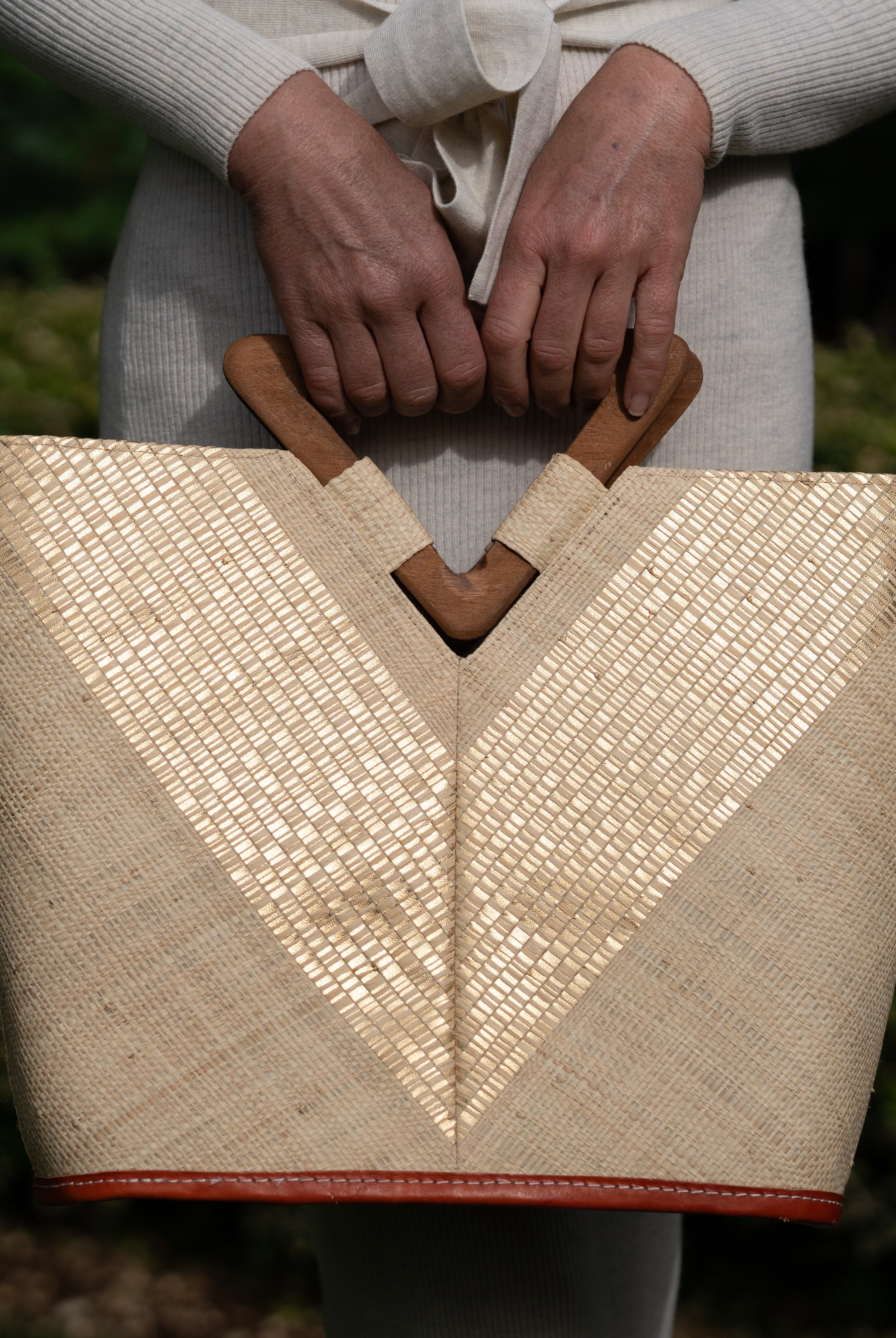 Model wearing Zuki Wynwood Natural Straw Handbag with Metallic Detailing & Wood Triangle Handle handmade loomed raffia palm fiber in a solid hue of natural straw color with incorporated vegan leather gold metallic strands in a V pattern across the front and back of the bag with assorted print African wax cloth liners and carved wooden triangle handle purse - Shebobo
