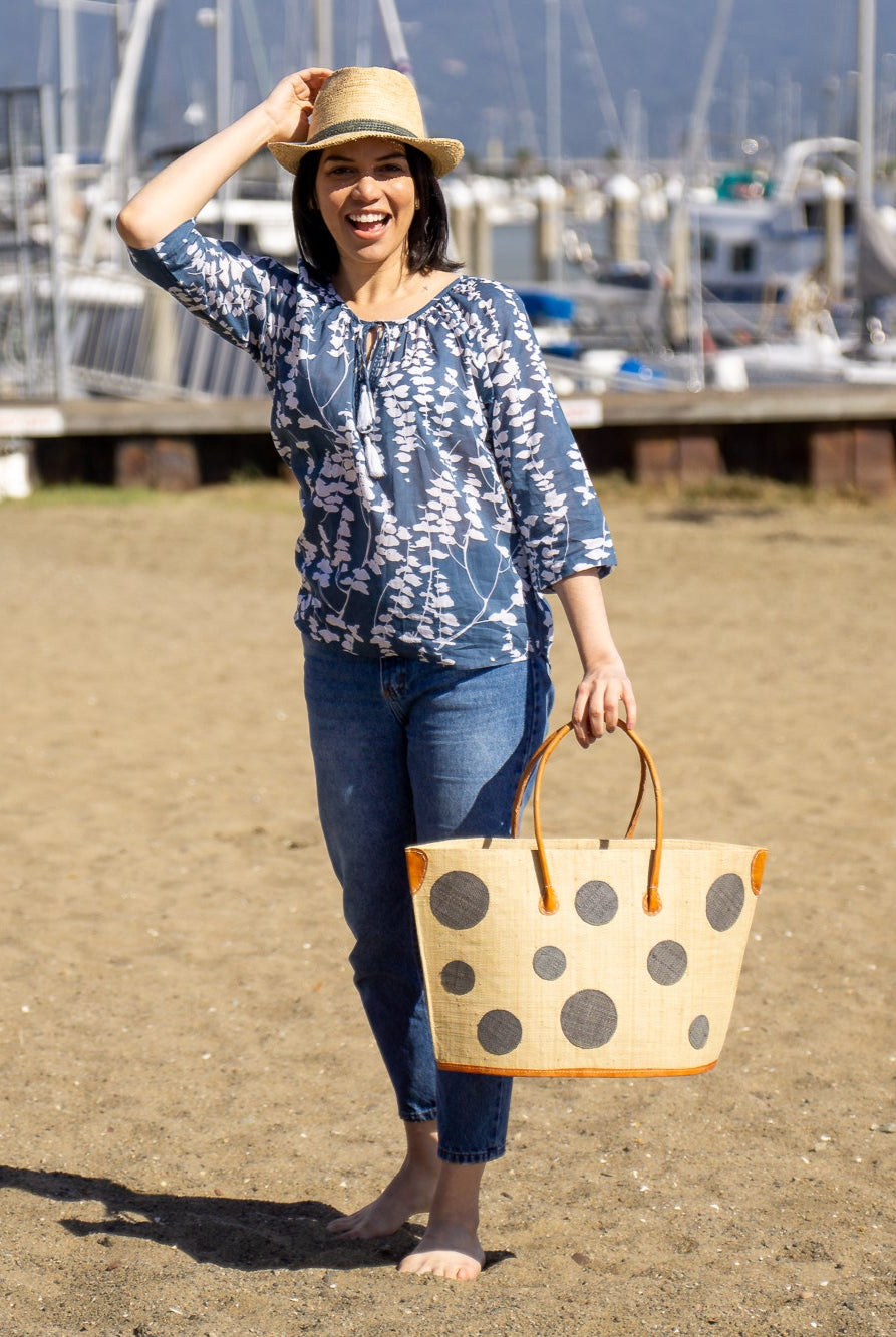 Model wearing Capri Polka Dot Grey Small Straw Tote Bag handmade loomed raffia palm fiber in a solid hue of natural straw color with various sized grey dots stitched on in a random pattern with leather handles and accents handbag shoulder bag beach bag - Shebobo (with Crochet Fedora Natural with Grey Band Unisex Straw Hat)
