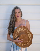 Model wearing 5.5" Brim Gigi Cinnamon and Natural Floral Embroidered Crochet Straw Sun Hats handmade crochet natural raffia palm fiber in a solid hue of cinnamon dark brown making a concentric pattern from the center top of the flattened crown spiraling out to the edge of the brim with matching adjustable raffia braid hat band and natural straw color floral pattern with leaves embroidered on the brim - Shebobo